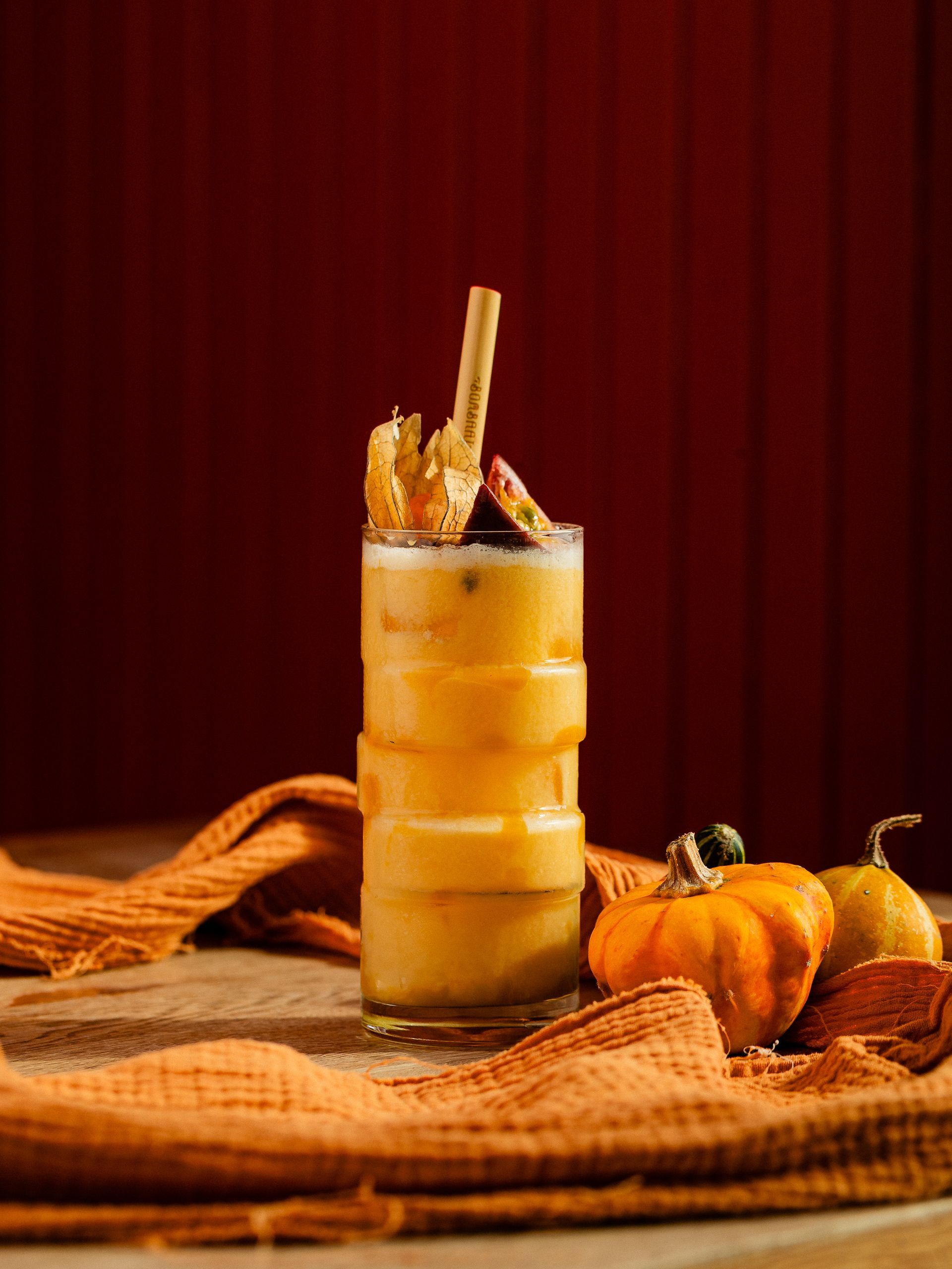 Hoge cocktail met laagjes sinaasappel, gegarneerd met passievrucht en een rietje. Pompoen- en sinaasappelstof op een houten ondergrond.