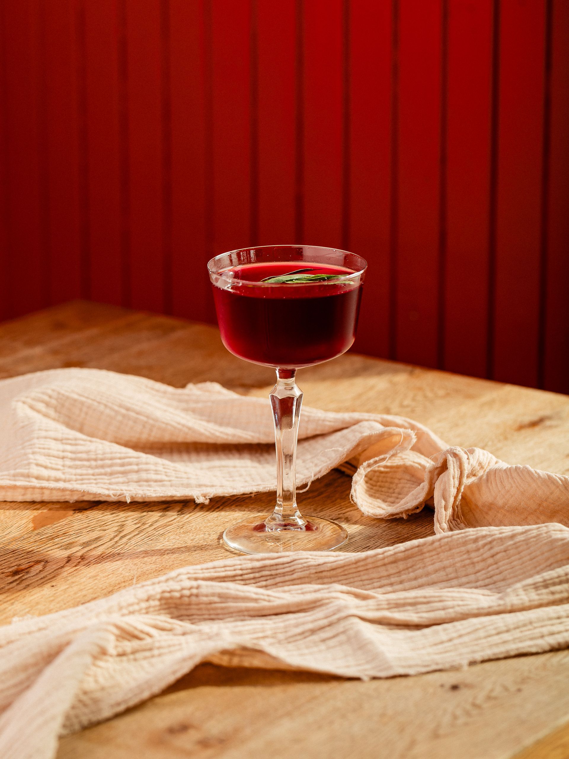 Een rode cocktail in een glas op voet, op een houten tafel bedekt met een beige tafelkleed.
