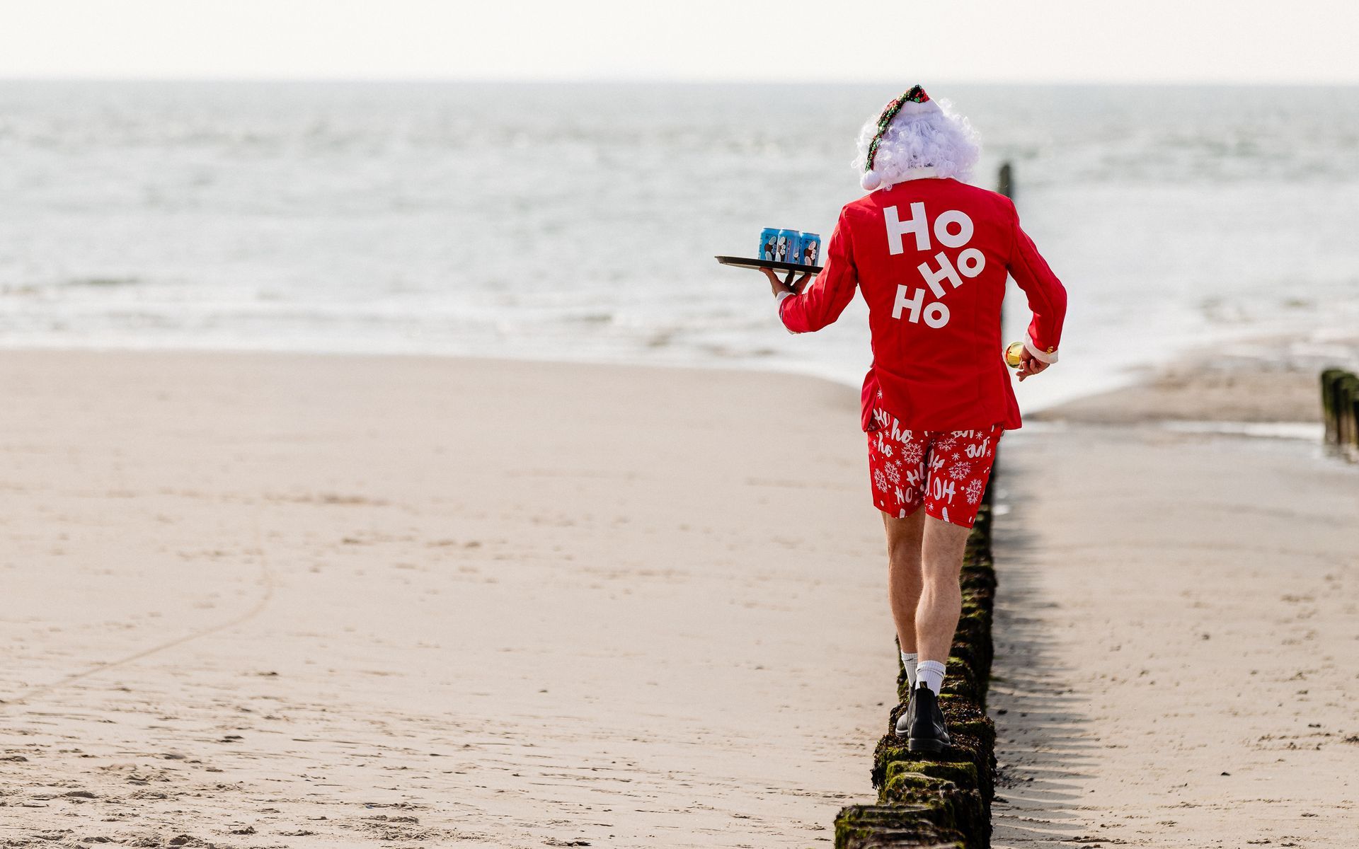 Een persoon verkleed als kerstman loopt met een dienblad over een houten pier op het strand. Hij draagt een rood pak met