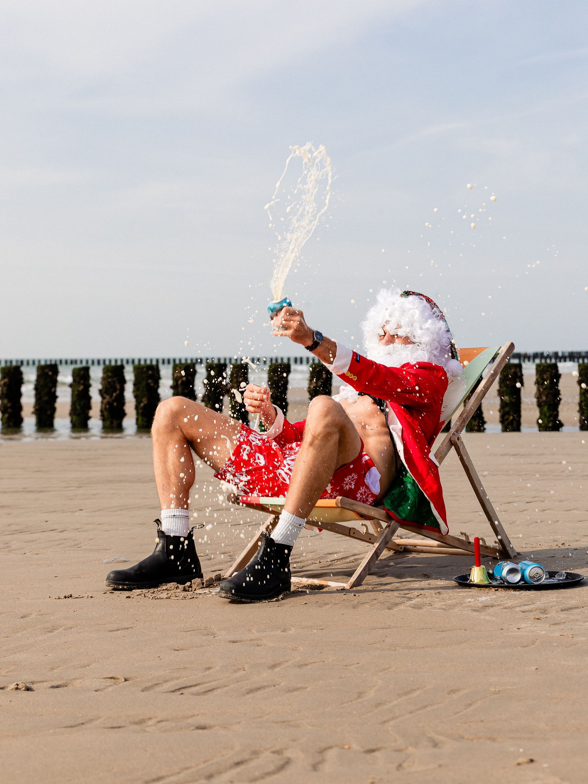 De kerstman zit in een strandstoel en spuit champagne op het zandstrand.