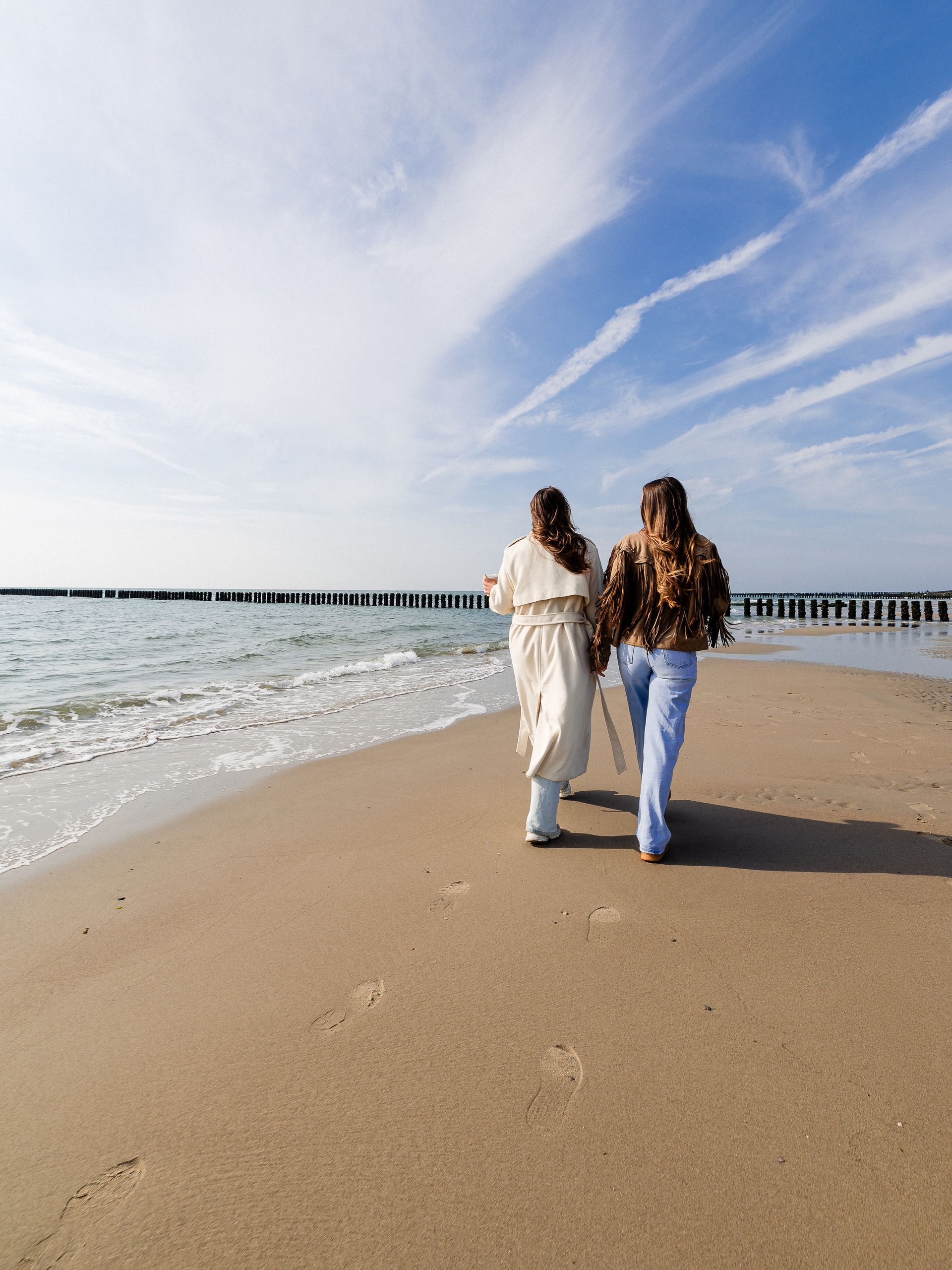 Twee mensen lopen over een zandstrand naast de oceaan, onder een blauwe hemel met wolken.