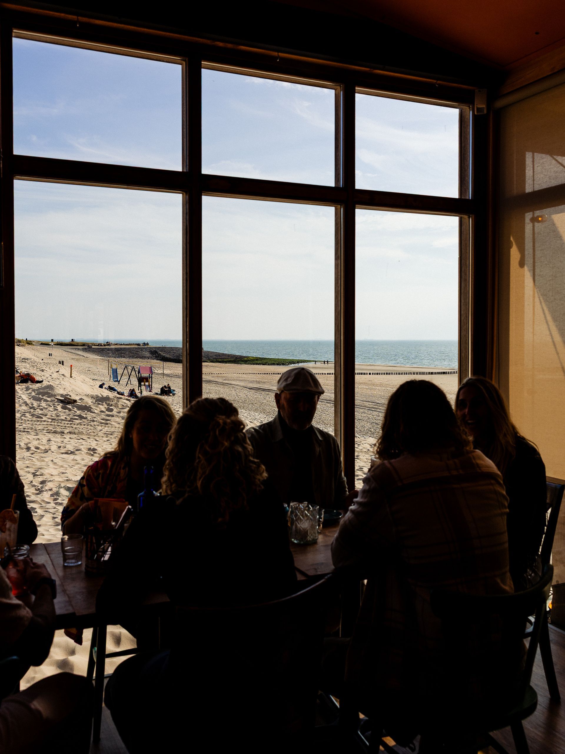 Silhouetten van mensen aan een tafel, groot raam biedt uitzicht op het strand en de oceaan.