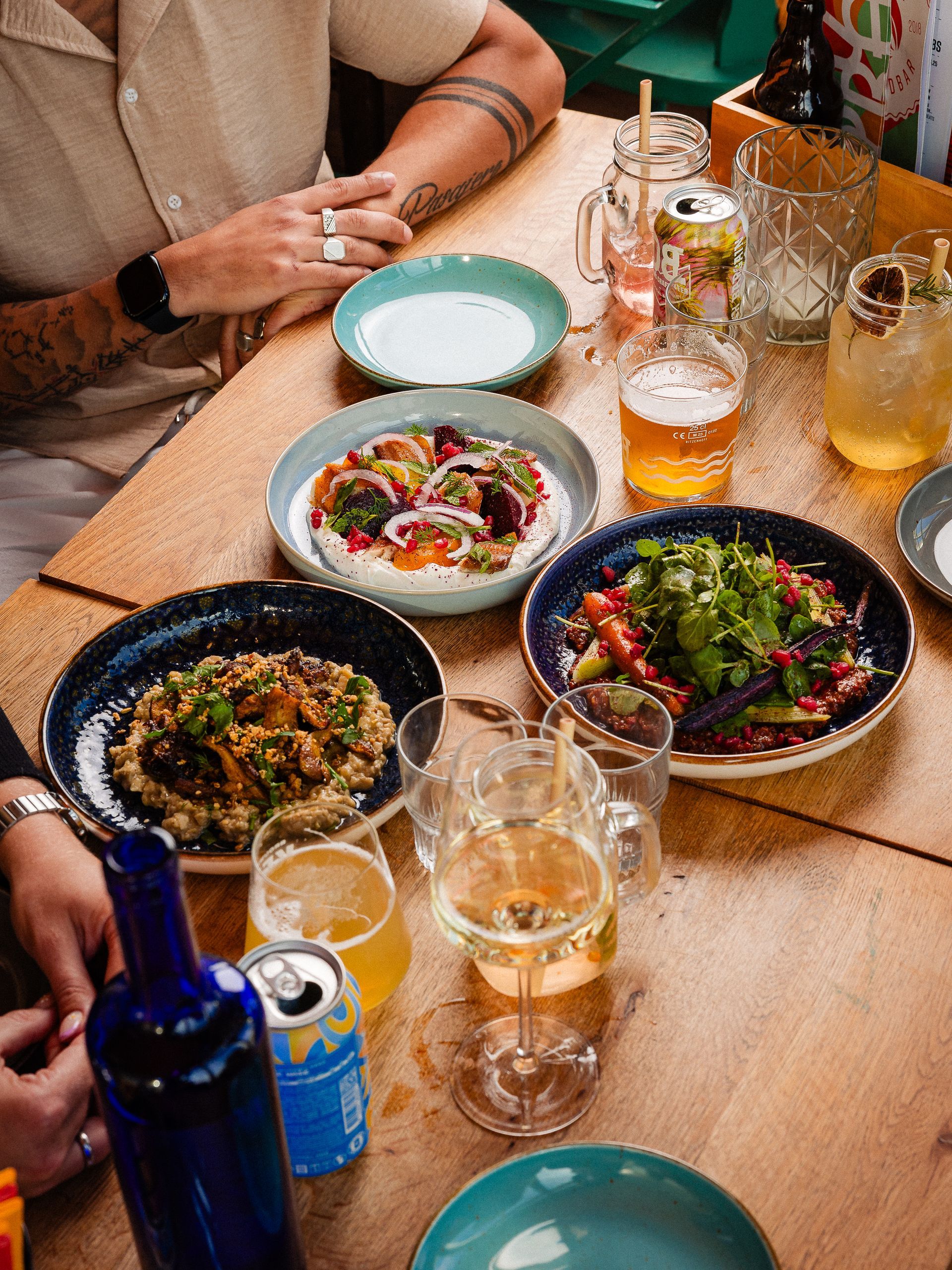 Tafel vol eten en drinken: gerechten uit het Midden-Oosten, bieren, wijnen en cocktails. Mensen zitten aan een houten tafel.