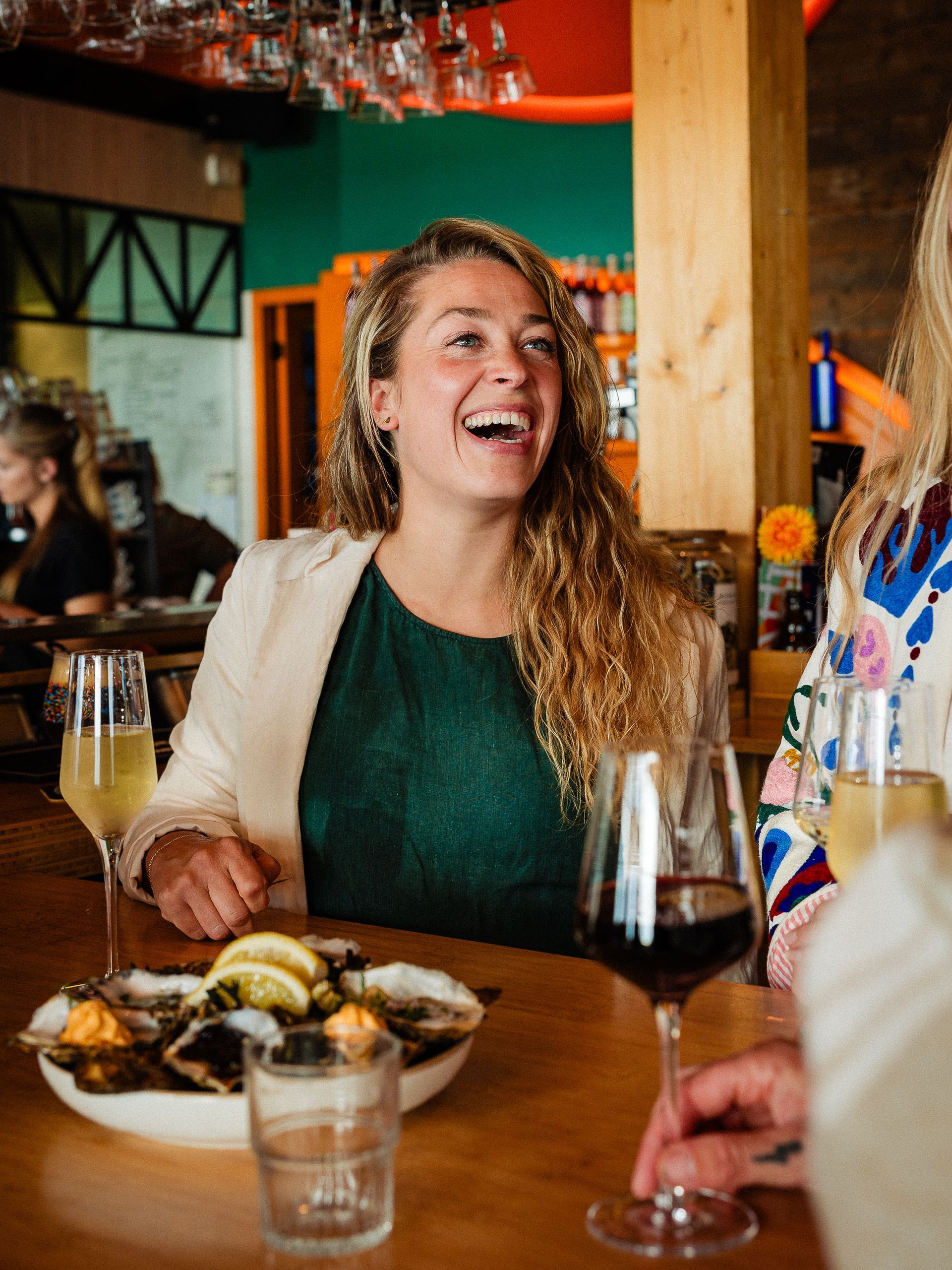 Een vrouw lacht aan de bar, terwijl ze een drankje met oesters vasthoudt en er iemand naast haar staat.