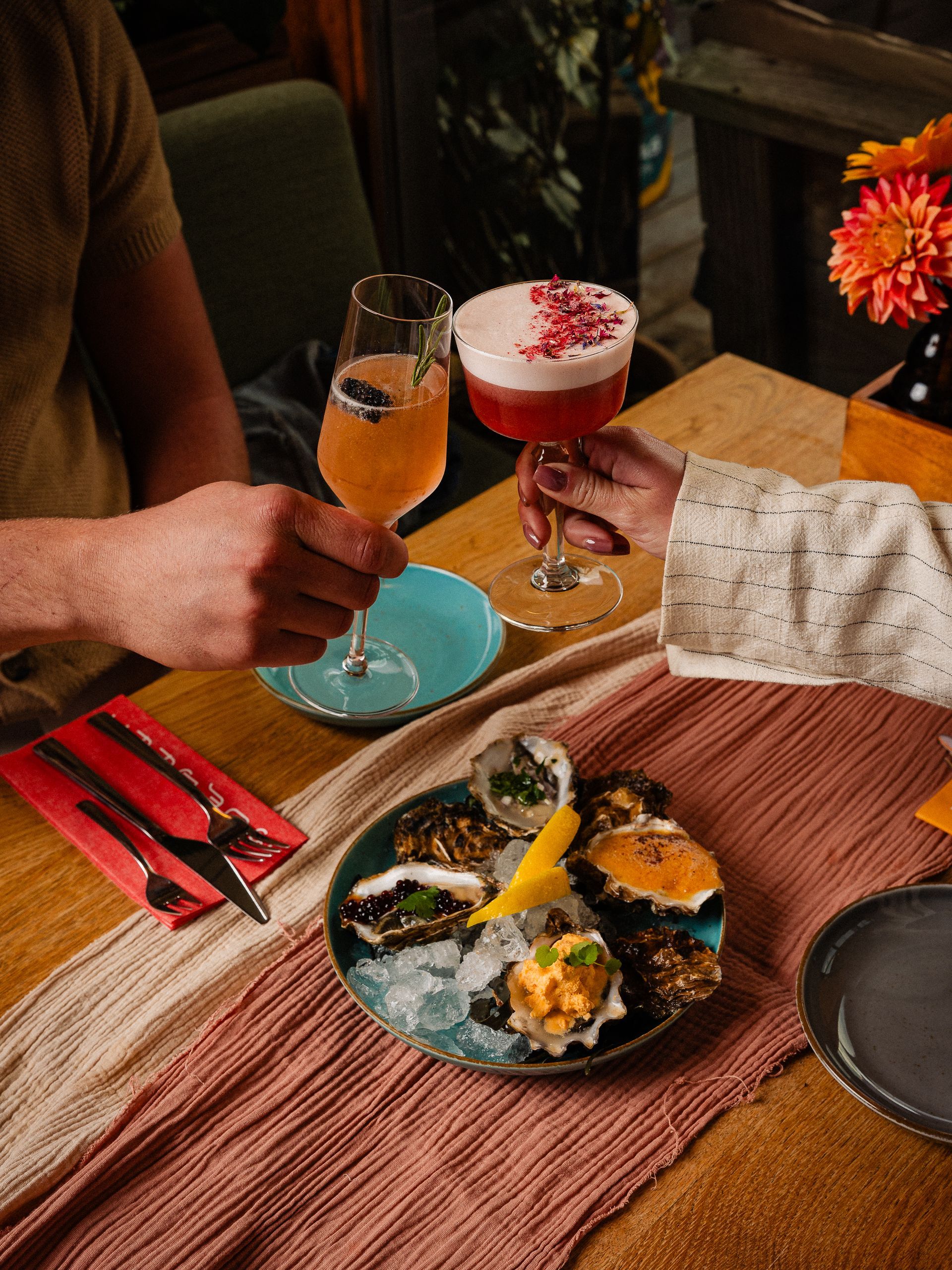 Twee mensen proosten met cocktails aan een tafel met oesters op een bord.