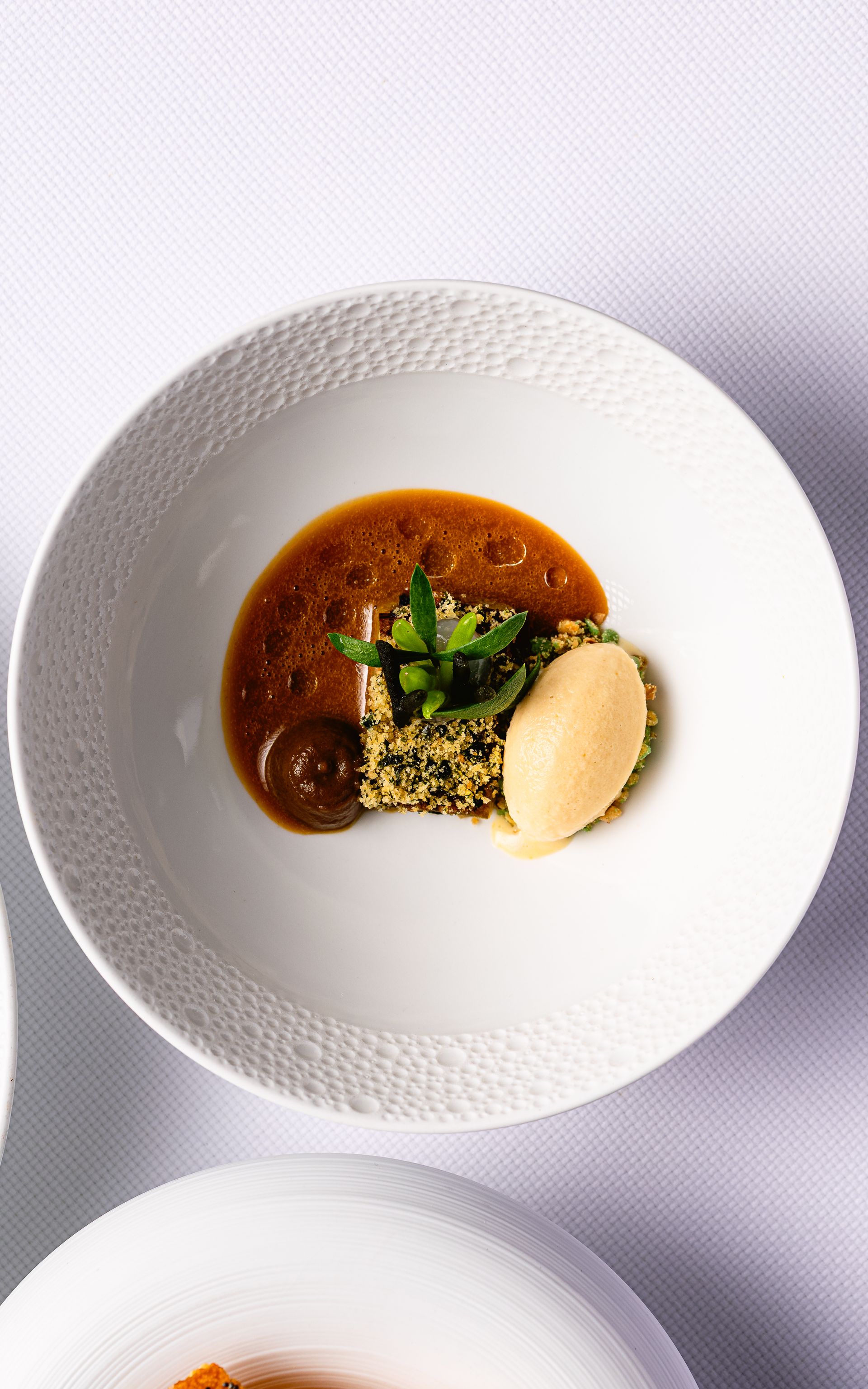 Witte kom met een gastronomisch gerecht: saus, groene kruiden, kruimels en een lichtbruine lepel.