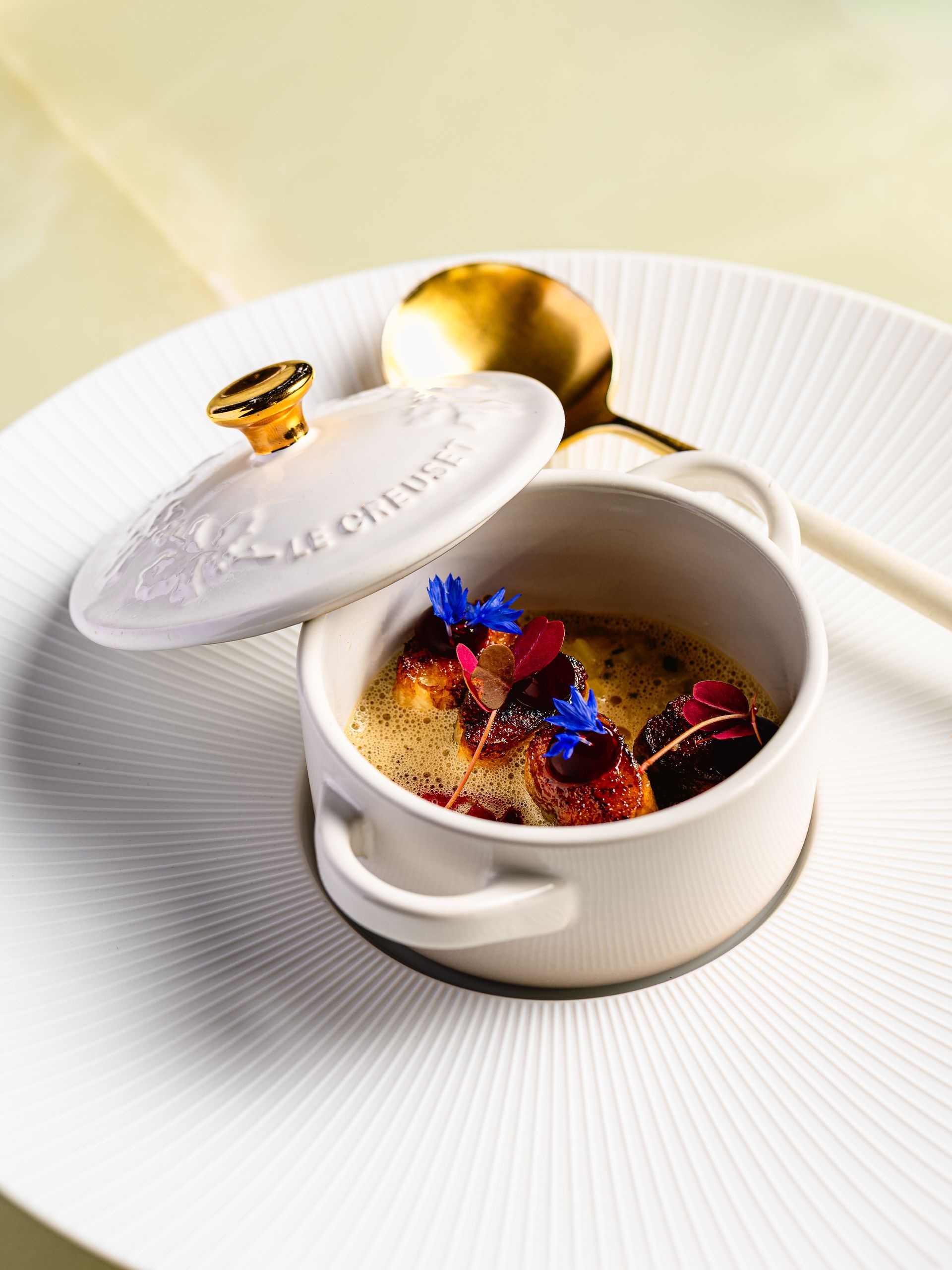 Klein wit potje met deksel, gevuld met eten, geserveerd op een wit bord met een gouden lepel.