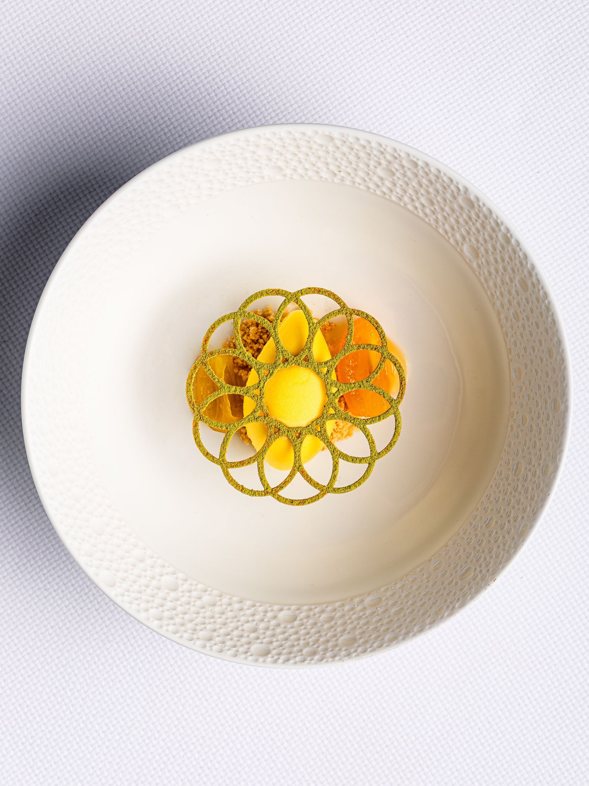 Geel dessert in een witte kom, versierd met een gouden, kantachtig patroon.