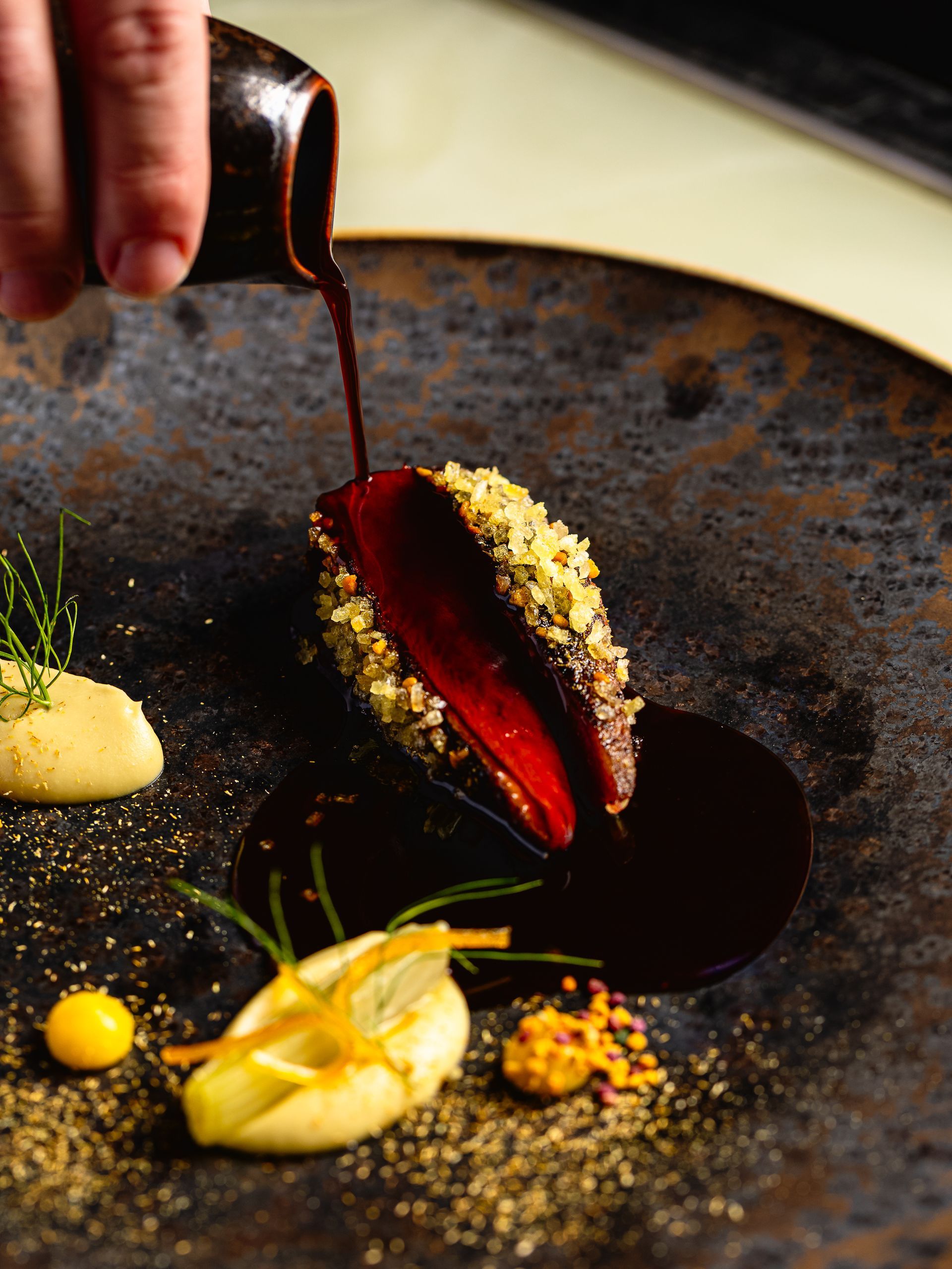 Een chef-kok giet een donkere saus over een prachtig opgemaakt gerecht met een gepaneerd stuk vlees in het midden.