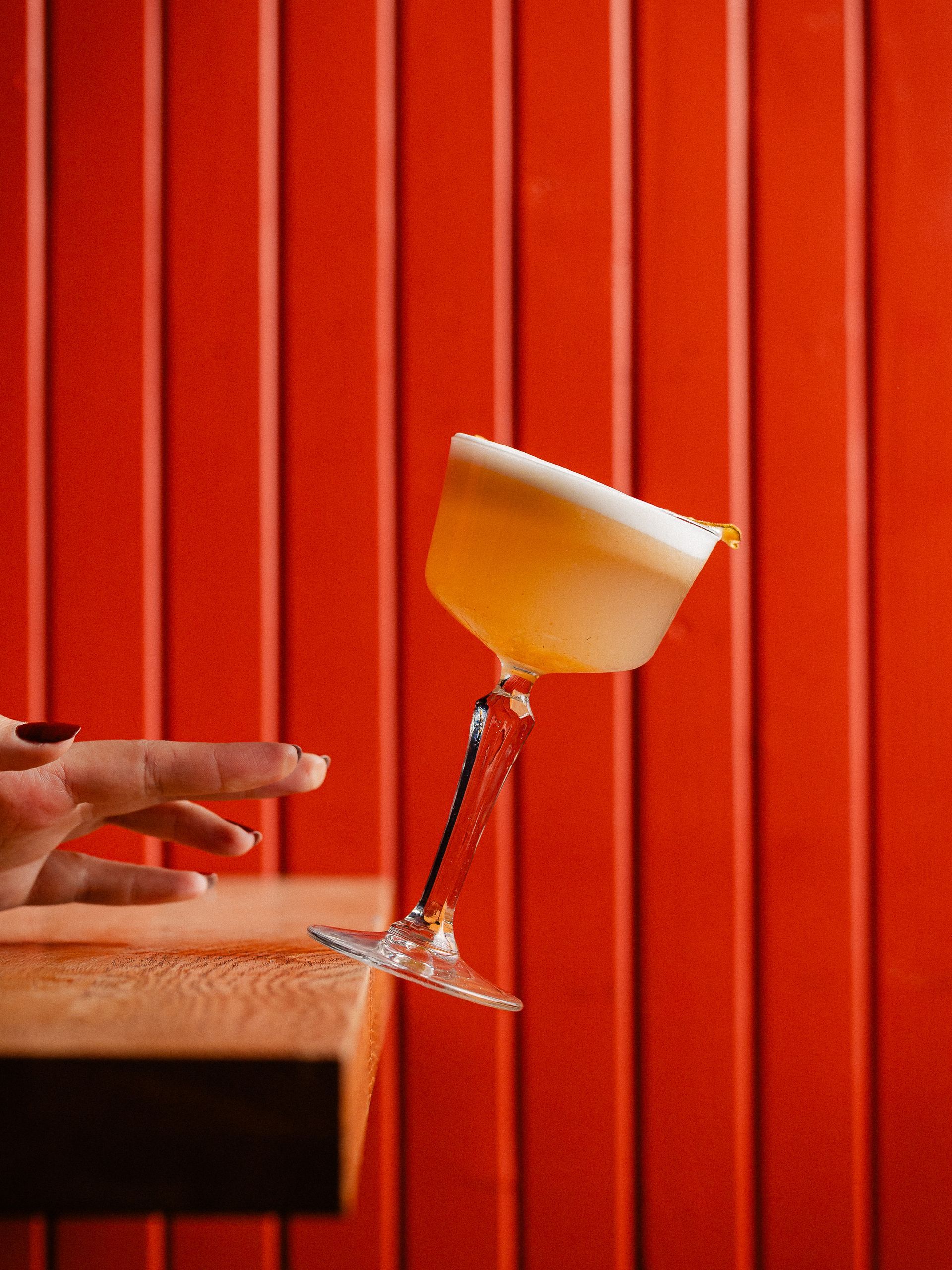 Een cocktailglas balanceert wankel op een houten rand, met een hand die uitsteekt tegen een rode lattenachtergrond.
