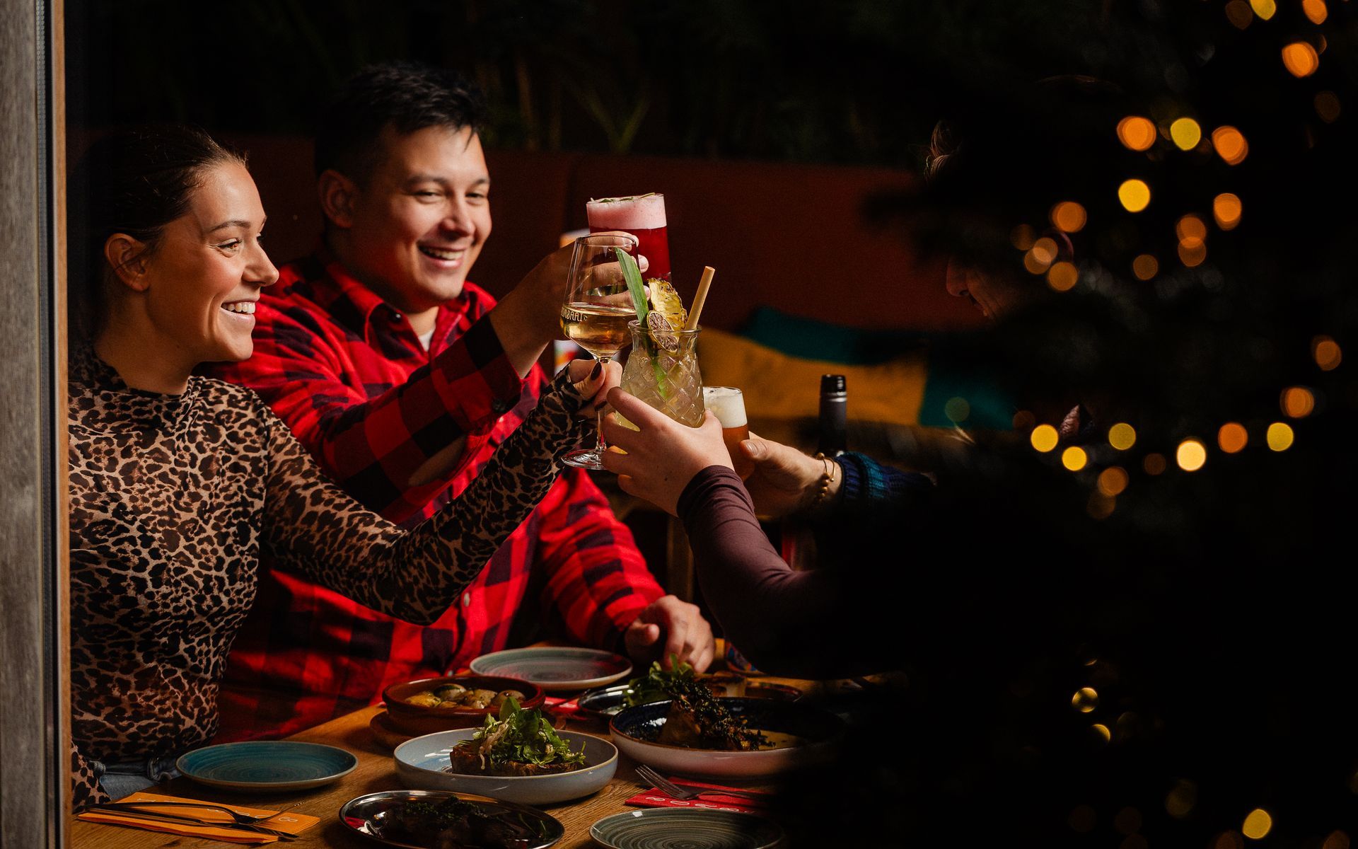 Mensen brengen een toast uit aan een tafel buiten. Kerstverlichting en eten zijn aanwezig.