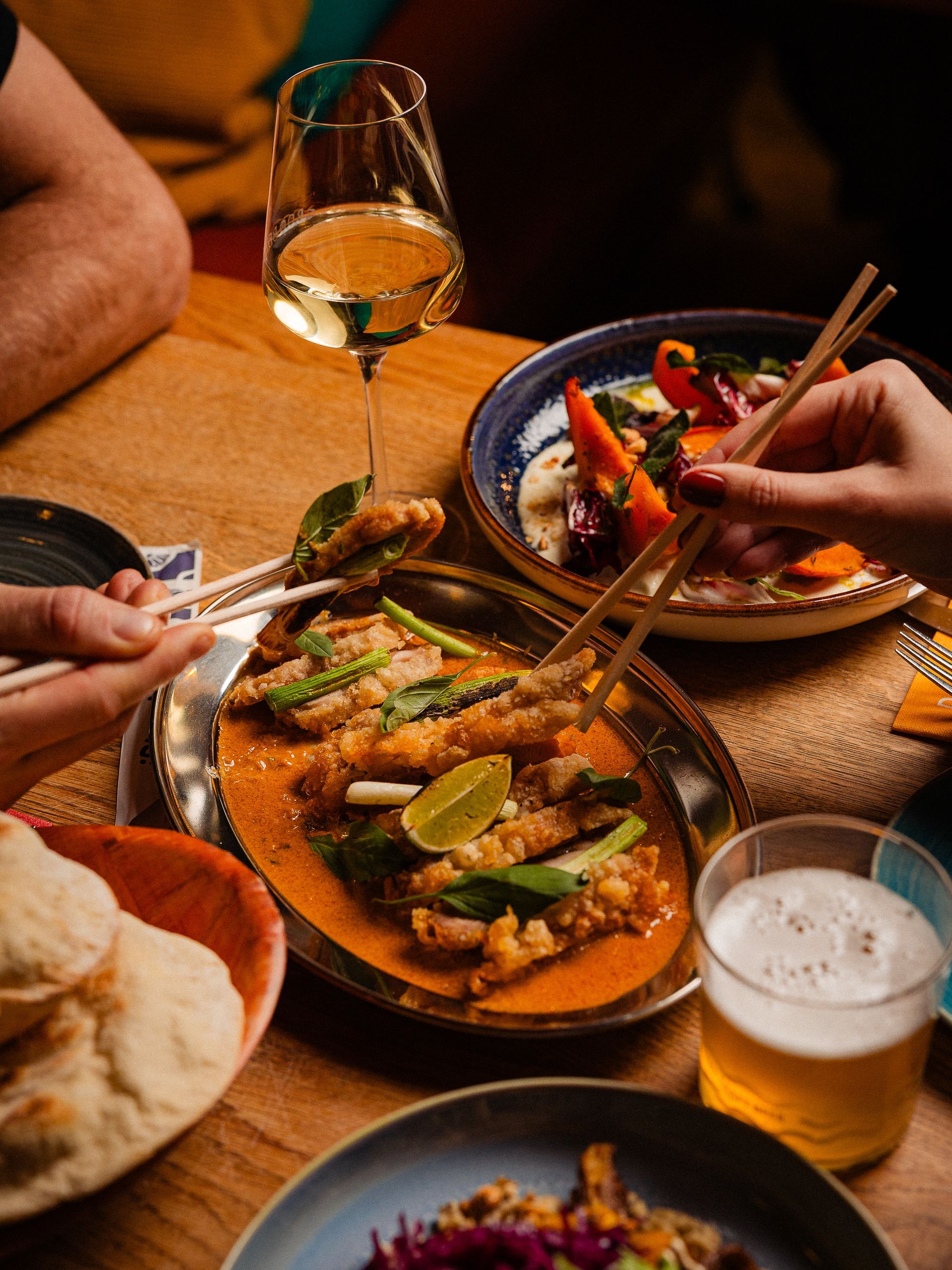 Mensen die met eetstokjes eten aan een tafel met borden vol eten, een glas wijn en een biertje.