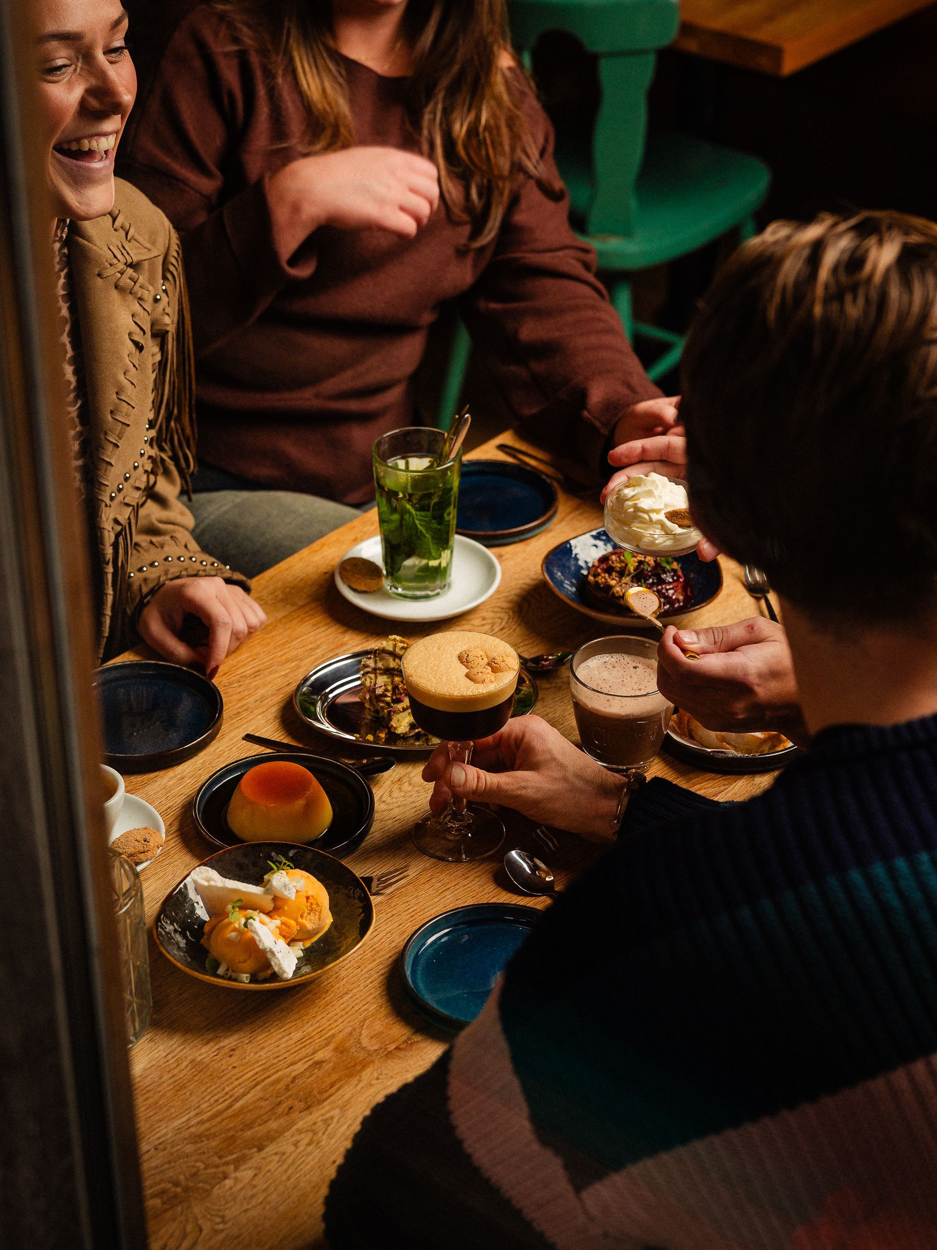 Mensen aan een tafel met desserts en drankjes, één van hen glimlacht. Donker interieur, houten tafel.