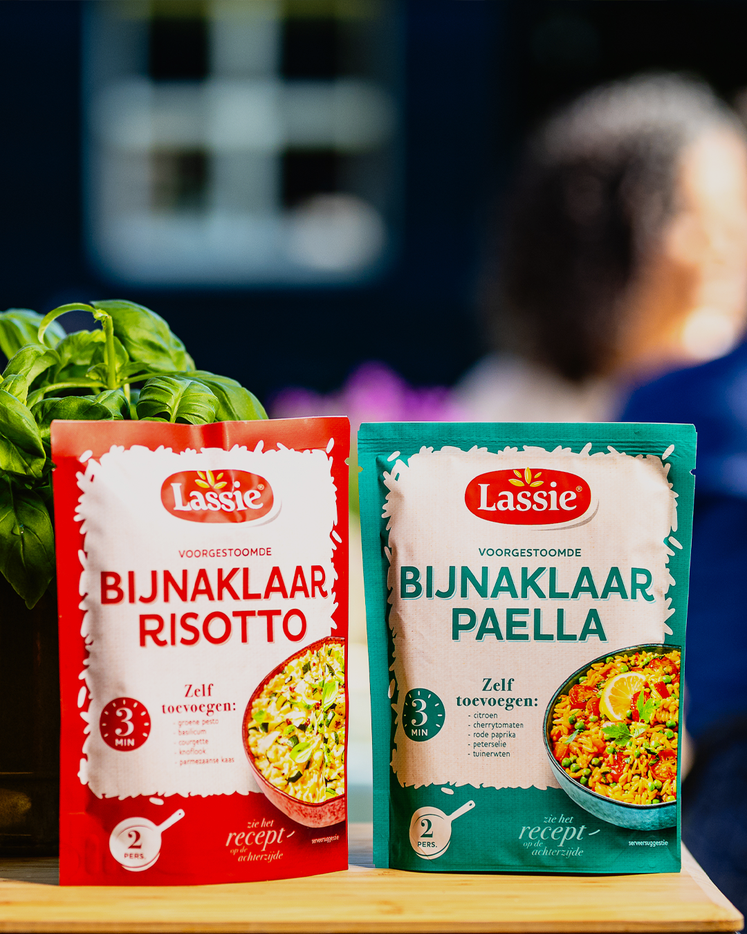 Twee pakjes kant-en-klare rijst van het merk Lassie: risotto (rood) en paella (groen), met op de achtergrond groen en een persoon.