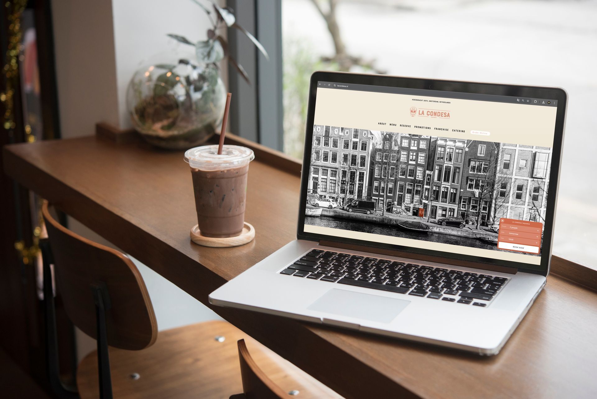 Een laptop met een webpagina, een kop ijskoffie en een stoel staan ​​op een houten vensterbank bij het raam.