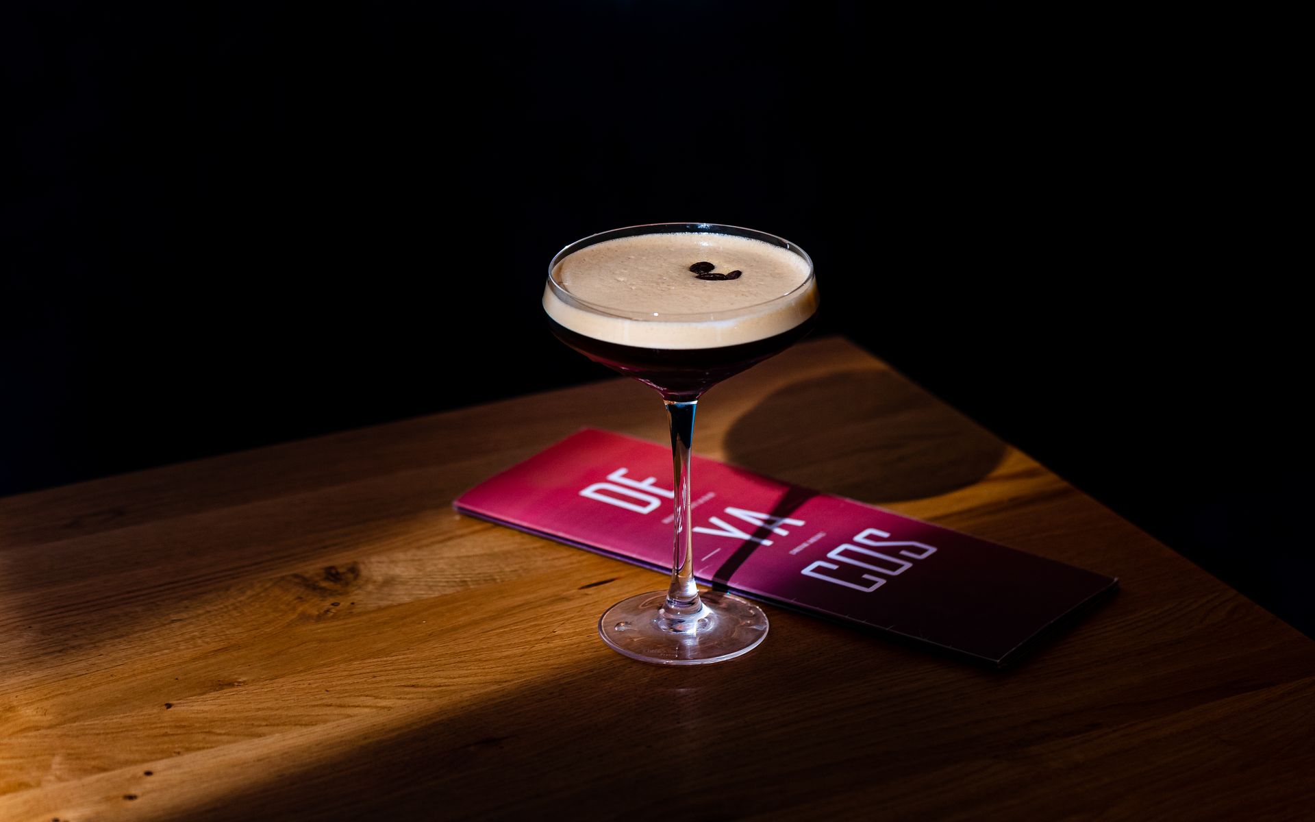 Espresso martini in een glas op voet, op een houten tafel, naast een rode menukaart, met een donkere achtergrond.