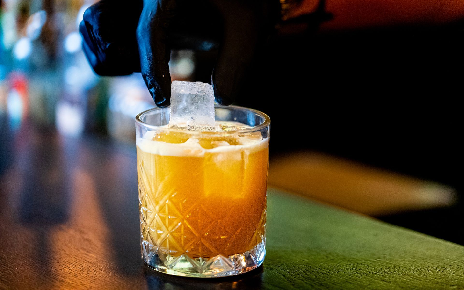 Een barman voegt een ijsblokje toe aan een cocktail in een glas met een patroon, dat op de bar staat.