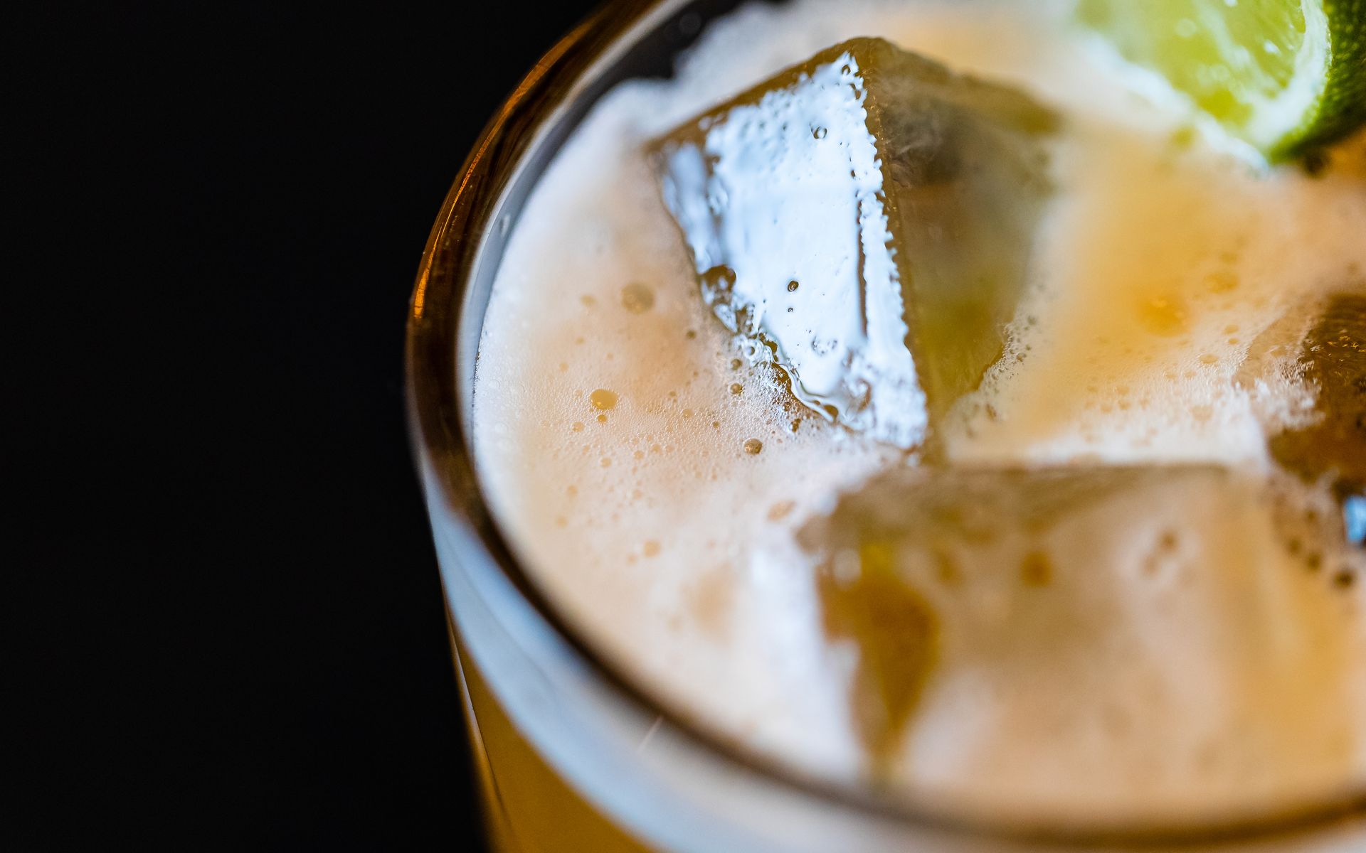 Close-up van een cocktail met ijsblokjes, schuim en een schijfje limoen in een glas tegen een donkere achtergrond.