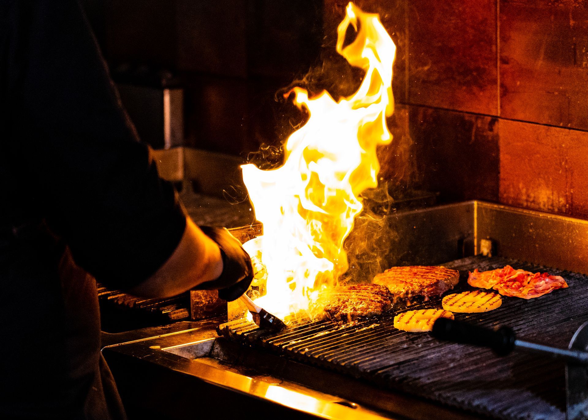 Vlammen omhullen de grill waarop hamburgers worden gebakken. Persoon in zwart shirt, donkere omgeving.