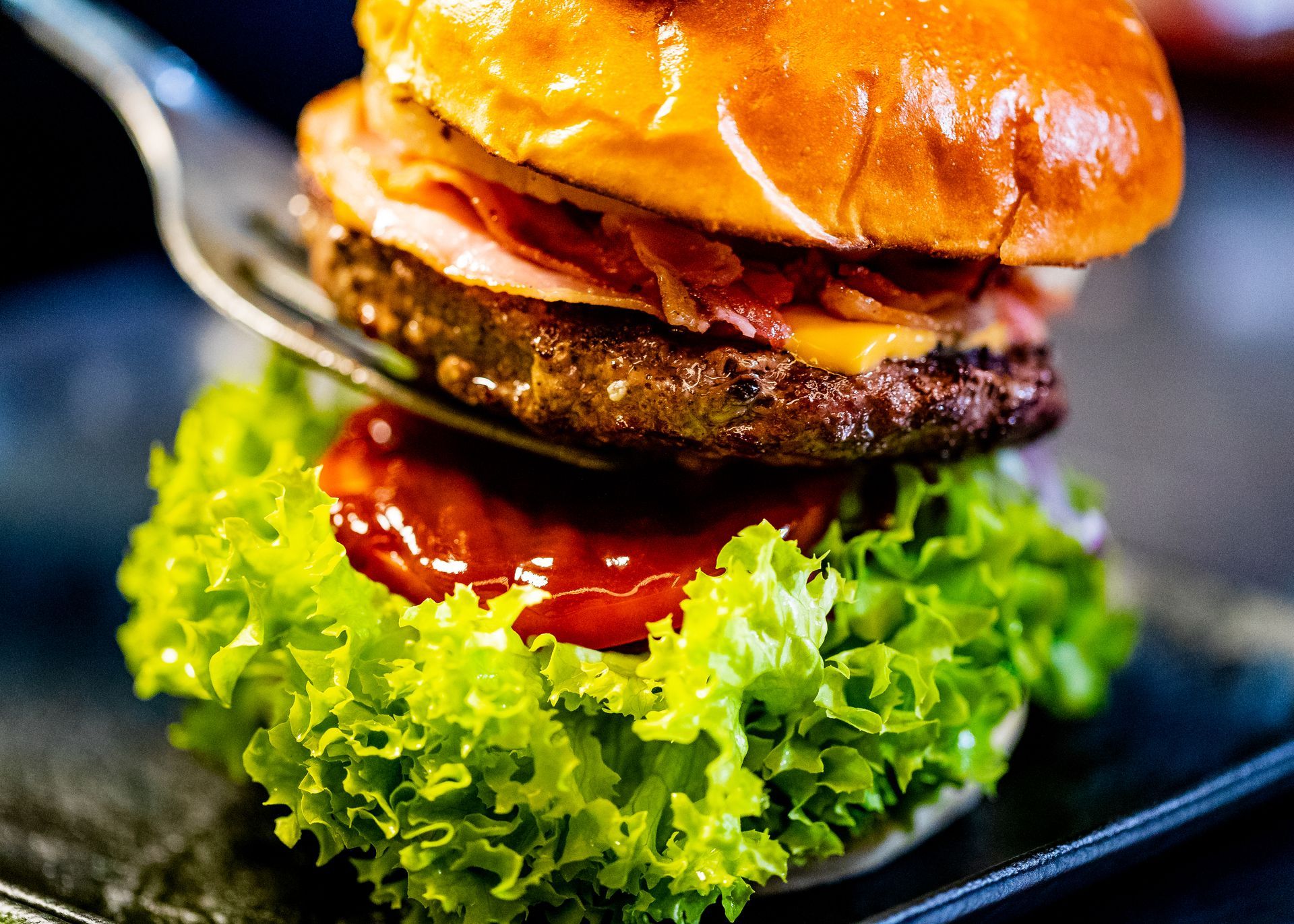 Een hamburger wordt in elkaar gezet: sla, hamburgerpatty, spek, kaas, broodje, en een vork houdt alles bij elkaar.