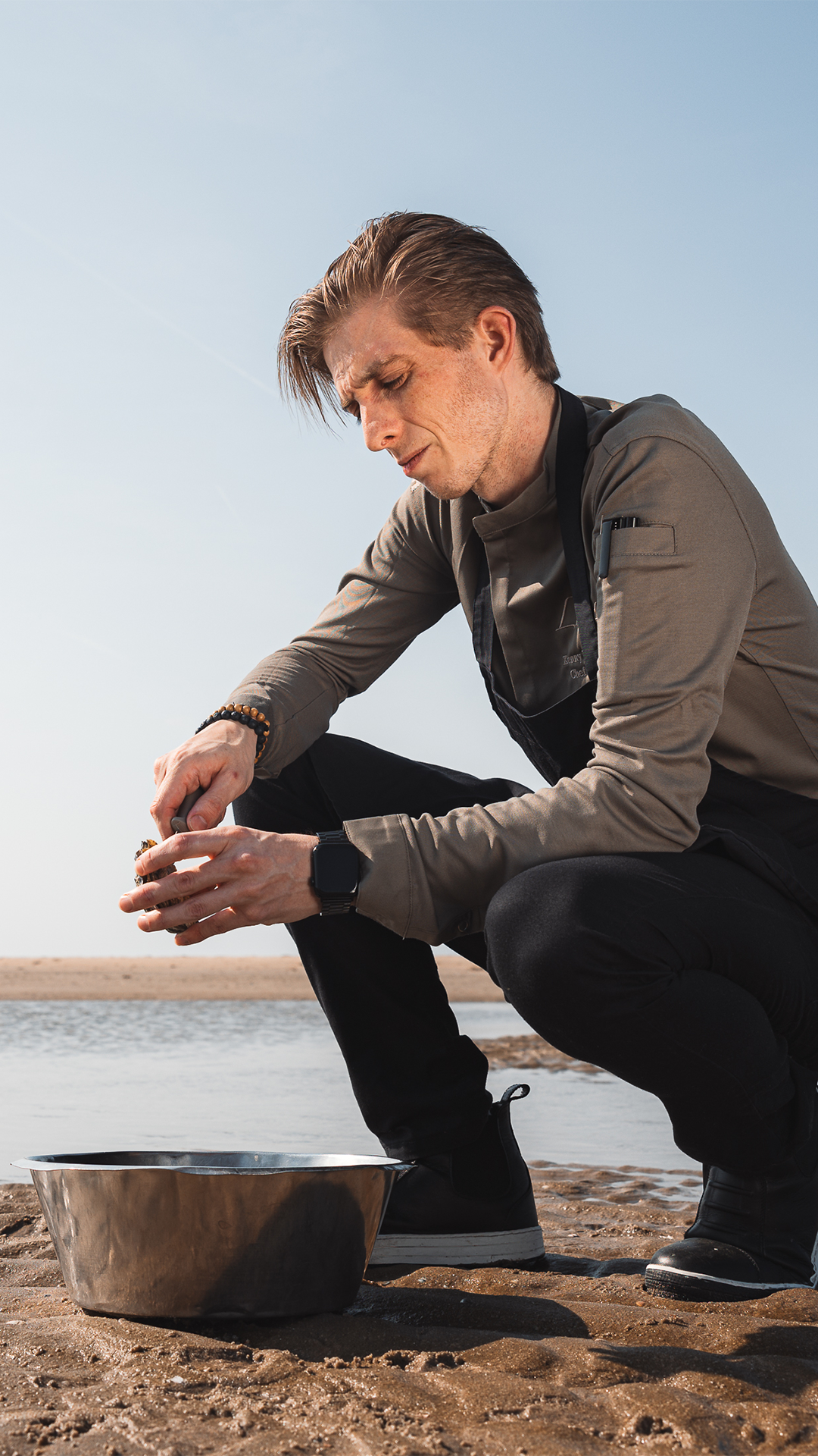 Een man hurkt op het strand en kijkt naar een voorwerp in zijn handen; een metalen kom staat ernaast en op de achtergrond is een watermassa te zien.