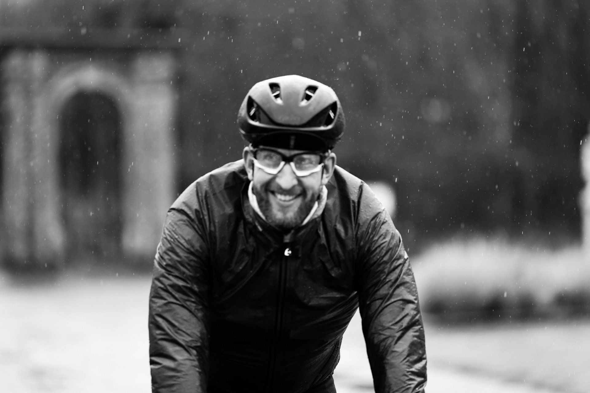 Een fietser met helm en bril lacht in de regen terwijl hij richting de camera fietst.