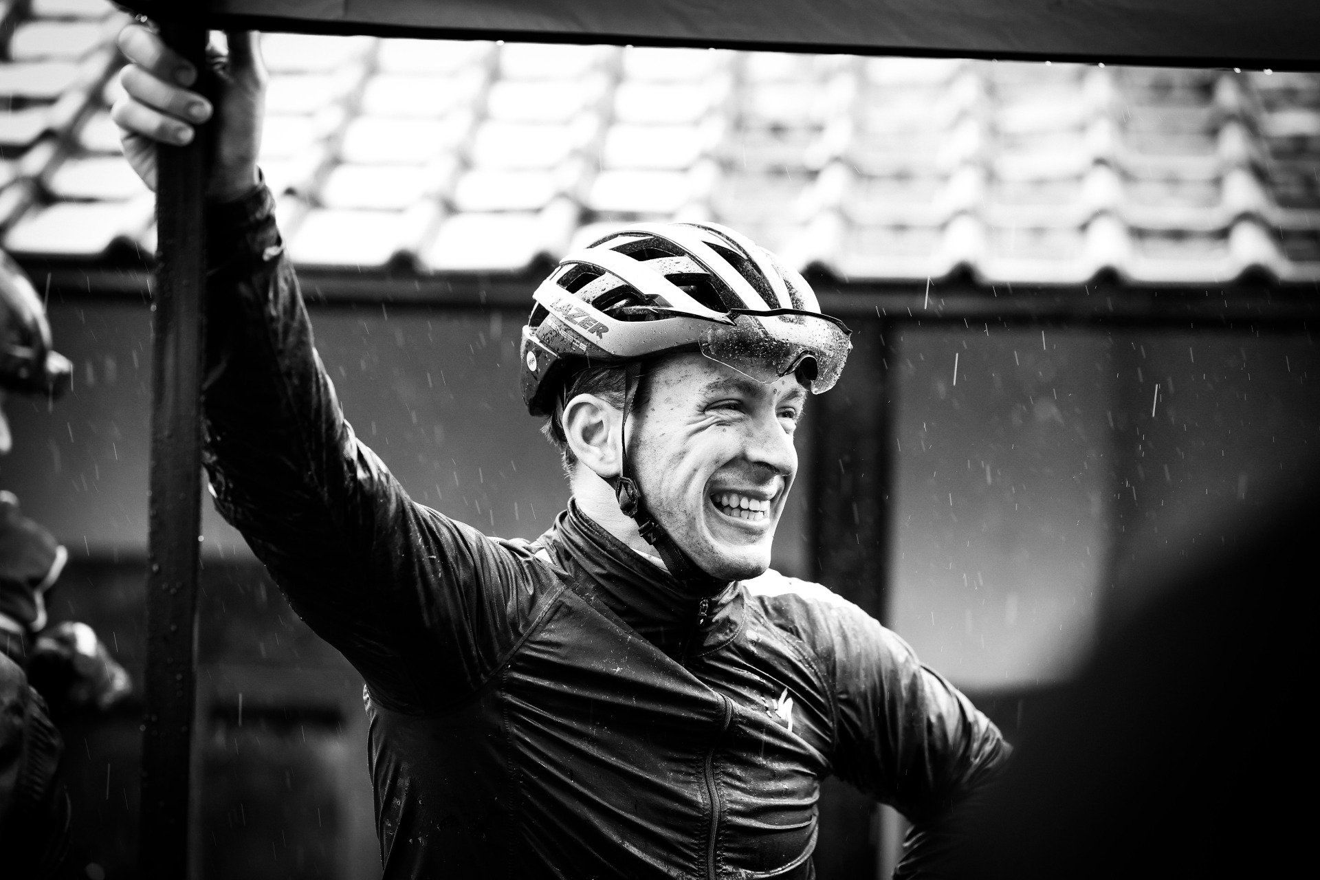 Een wielrenner steekt triomfantelijk zijn arm omhoog, lacht, draagt een helm en een natte jas, terwijl de regen valt.