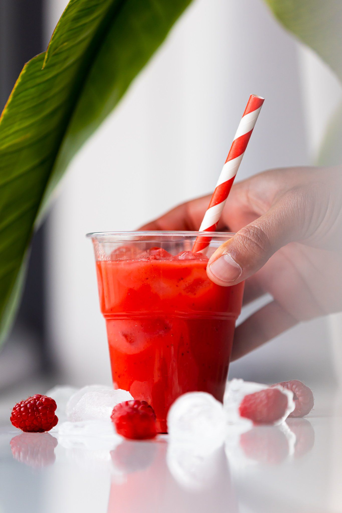 Een rode smoothie in een doorzichtige beker met een gestreept rietje, vastgehouden door iemands hand, omringd door ijsblokjes en frambozen.