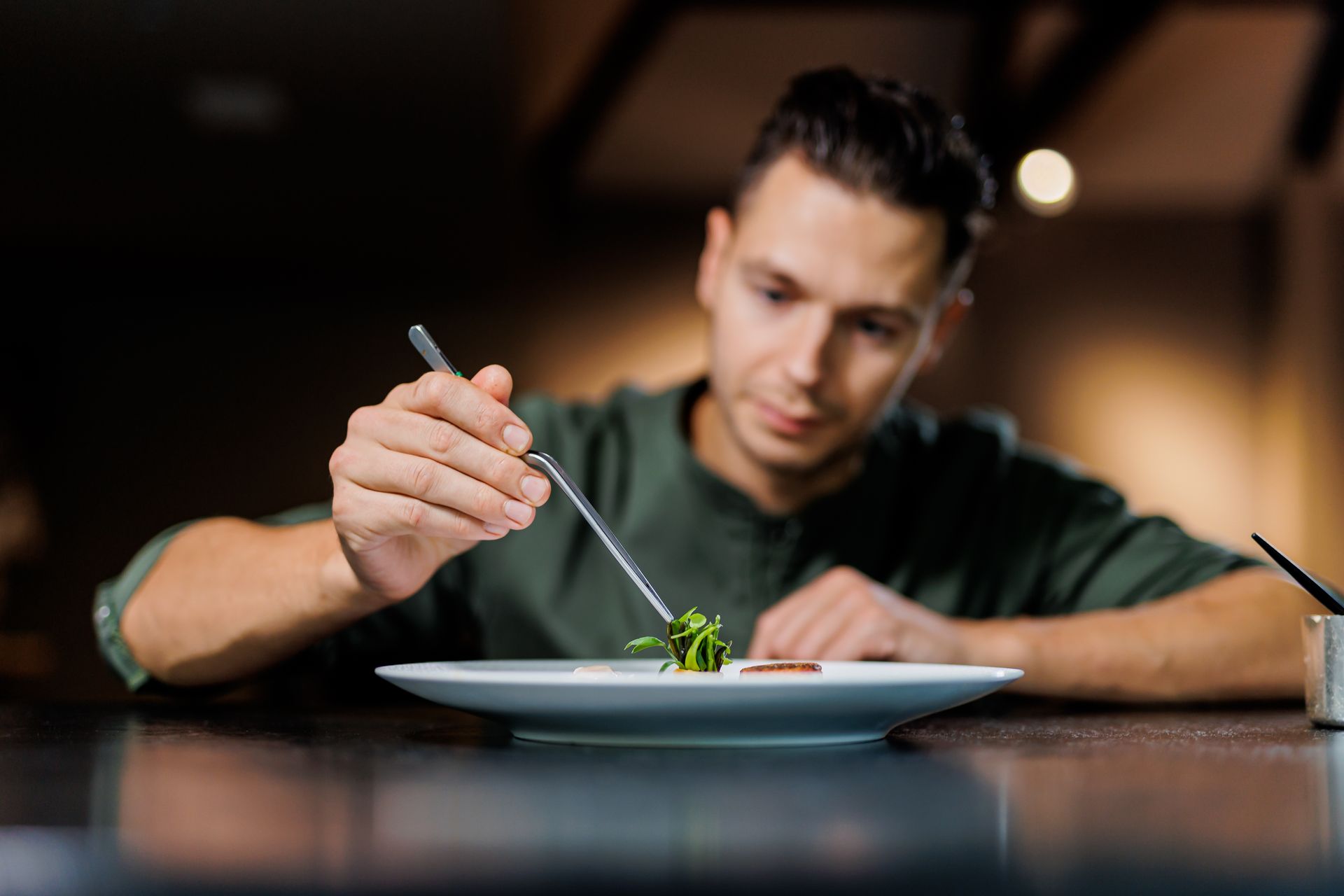 De chef-kok garneert een gerecht met een pincet op een wit bord aan een donkere toonbank.