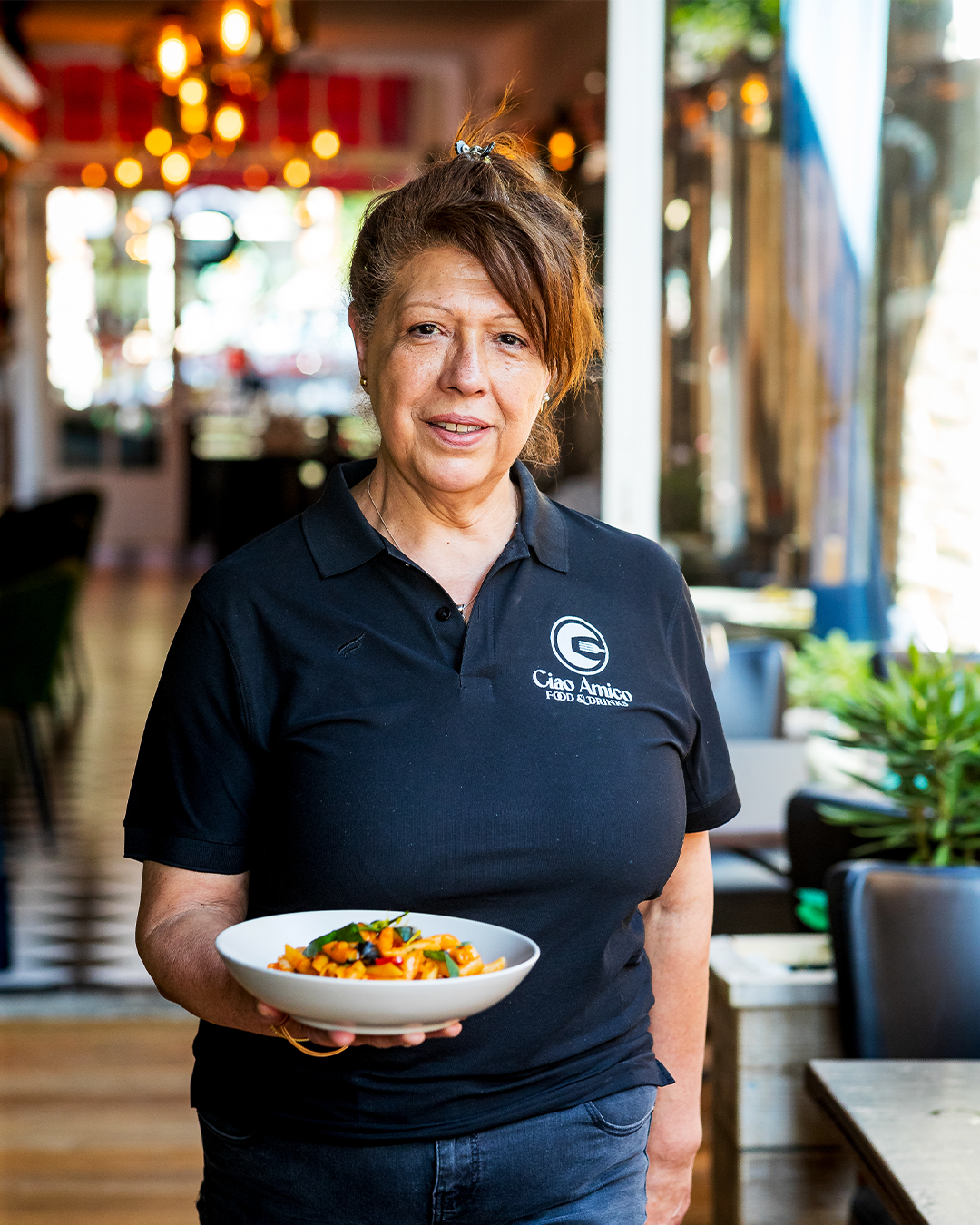 Vrouw met een bord pasta in haar handen, gekleed in een zwart shirt met logo, staat in een restaurant.