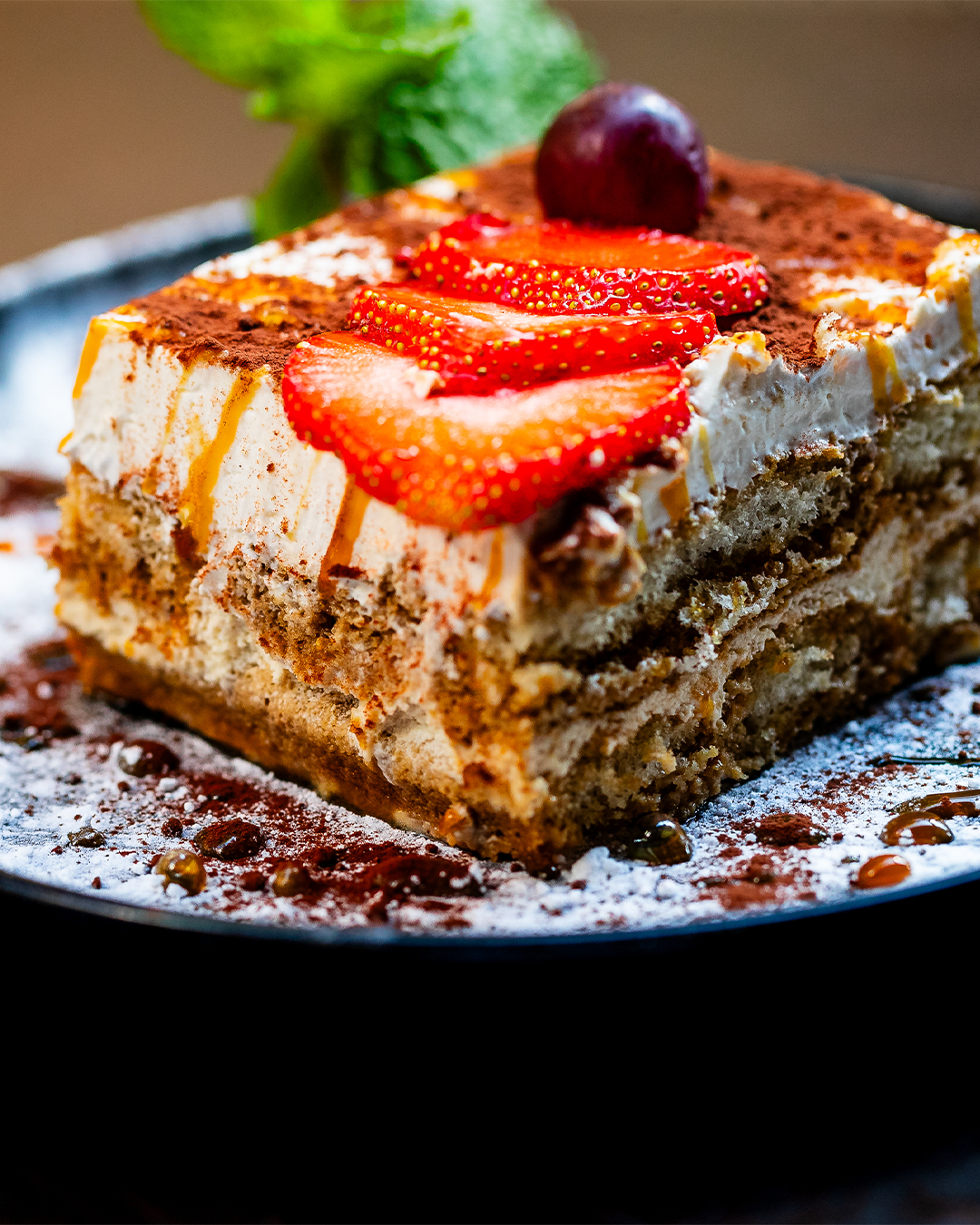 Een plakje tiramisu met aardbeien, een druif en munt op een bord, bestrooid met cacao en poedersuiker.