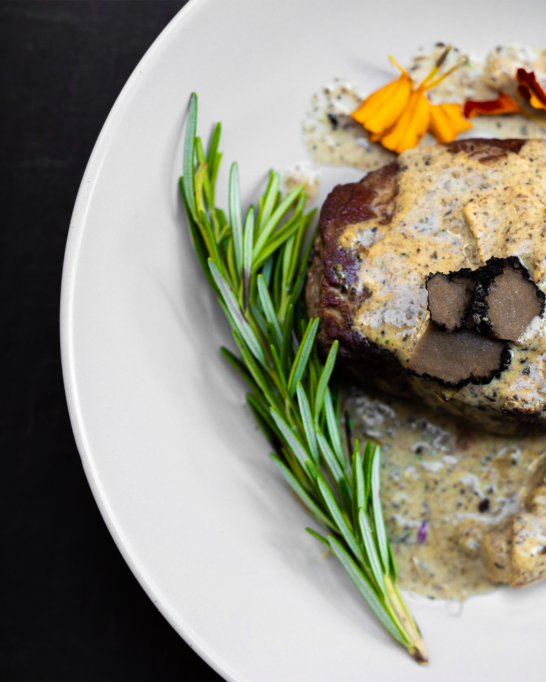Biefstuk met truffelsaus en een takje rozemarijn op een wit bord, zwarte achtergrond.