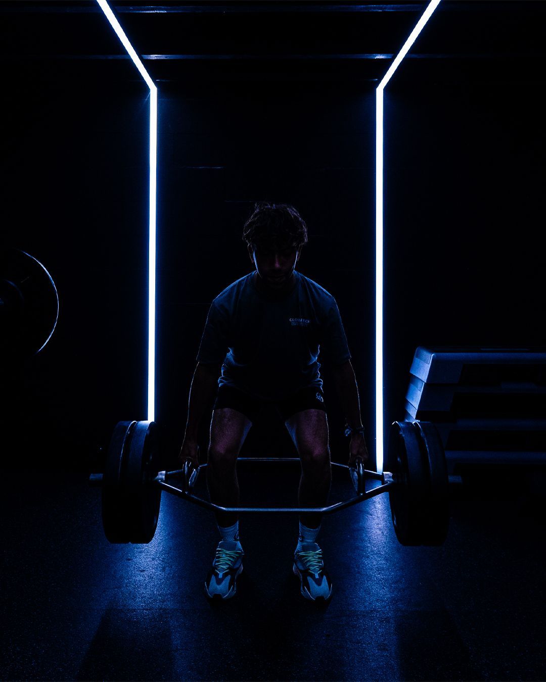 Silhouet van een persoon die een zeshoekige halterstang optilt in een donkere sportschool met blauwe neonverlichting.