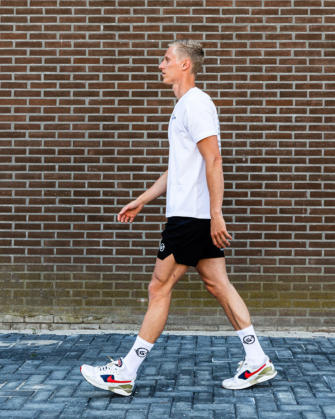 Een man in een wit shirt en een zwarte korte broek loopt over een met bakstenen bestrate stoep voor een bakstenen muur.