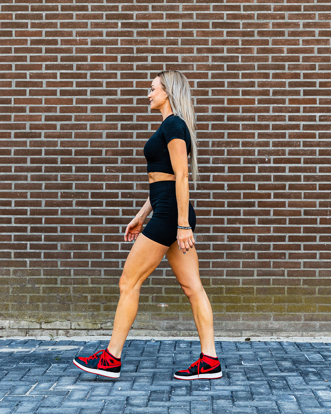 Vrouw in zwarte sportkleding, rode sneakers, lopend, met een bakstenen muur op de achtergrond.