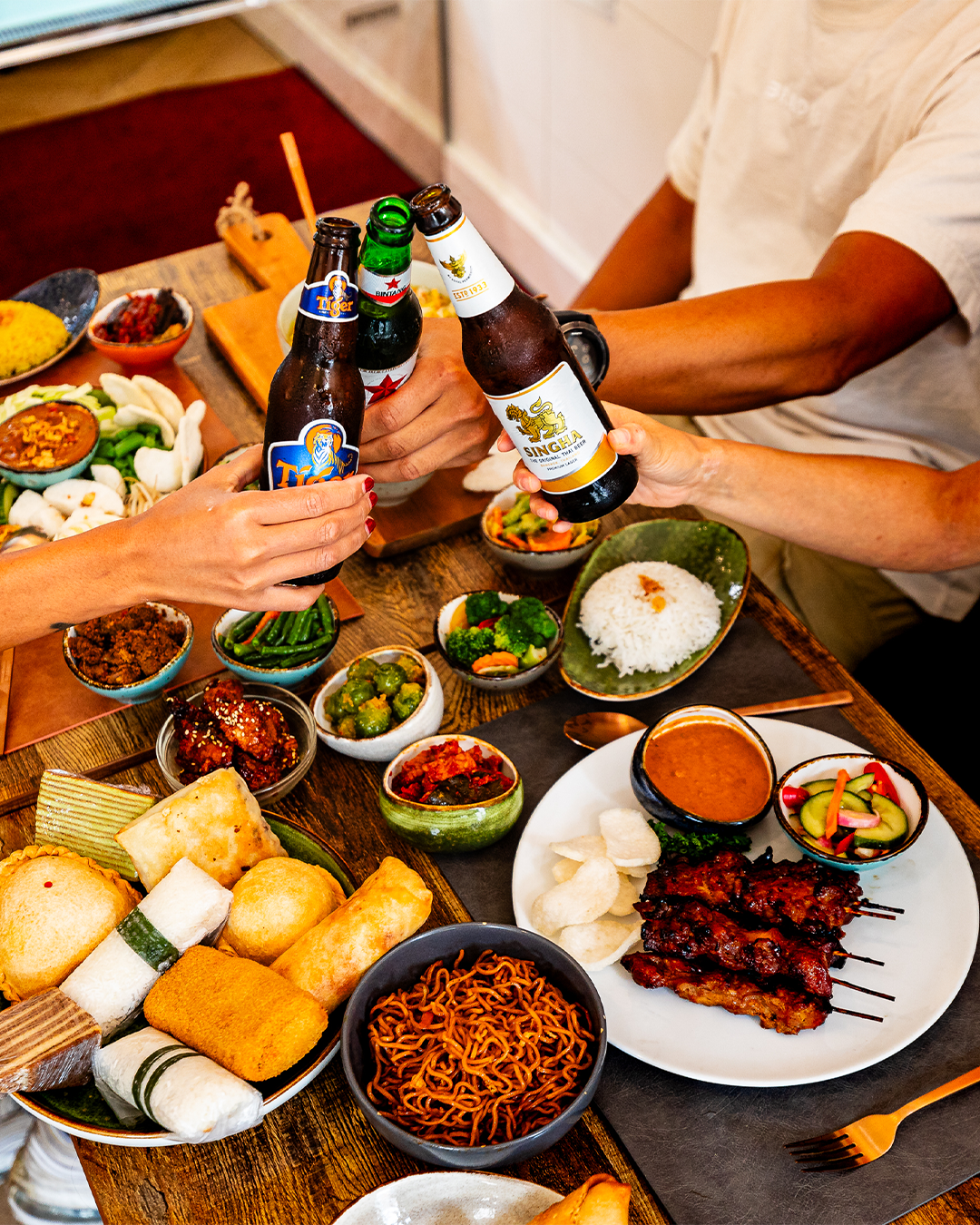 Mensen brengen een toast uit op bierflesjes boven een tafel vol Indonesisch eten.