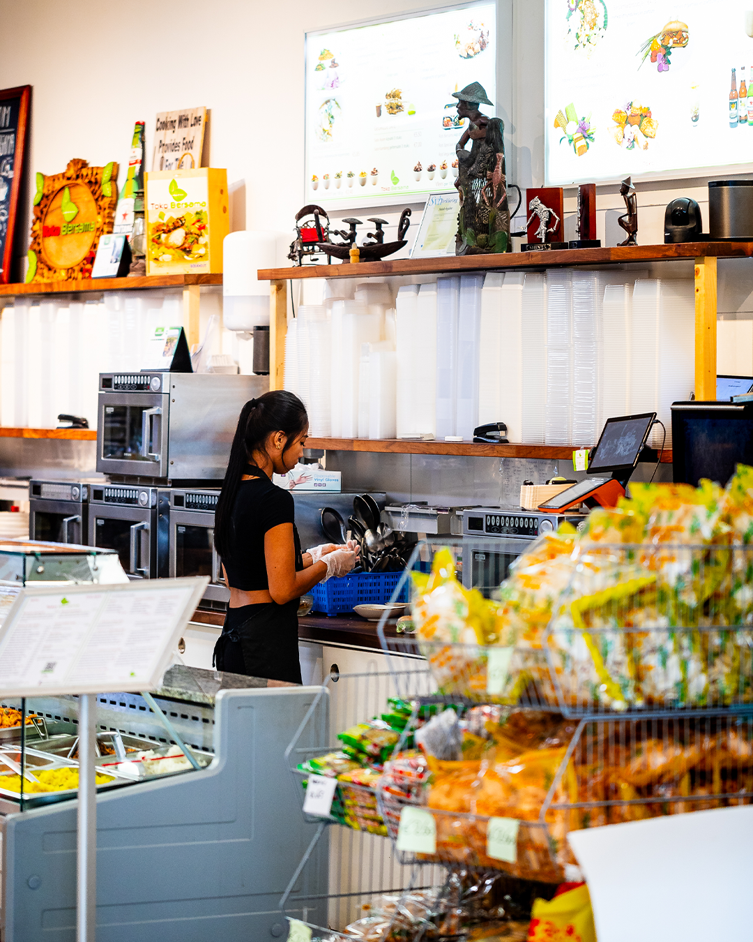 Vrouw werkt achter een toonbank in een winkel met schappen vol koopwaar en een vitrine.