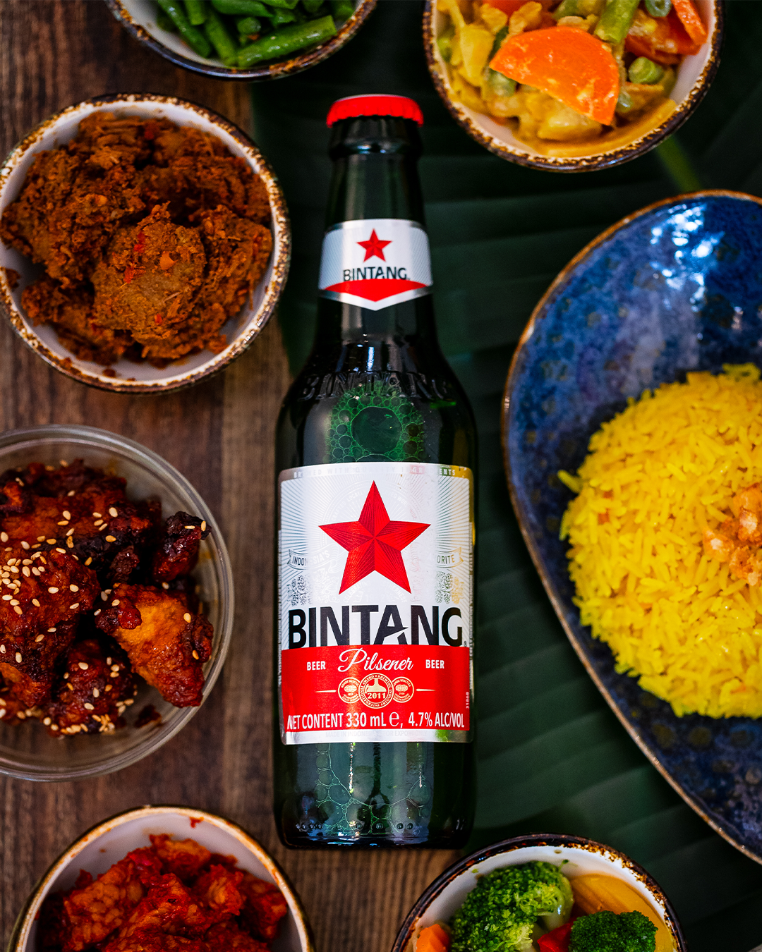 Een fles Bintang-bier omringd door schalen met Indonesisch eten op een groen blad.