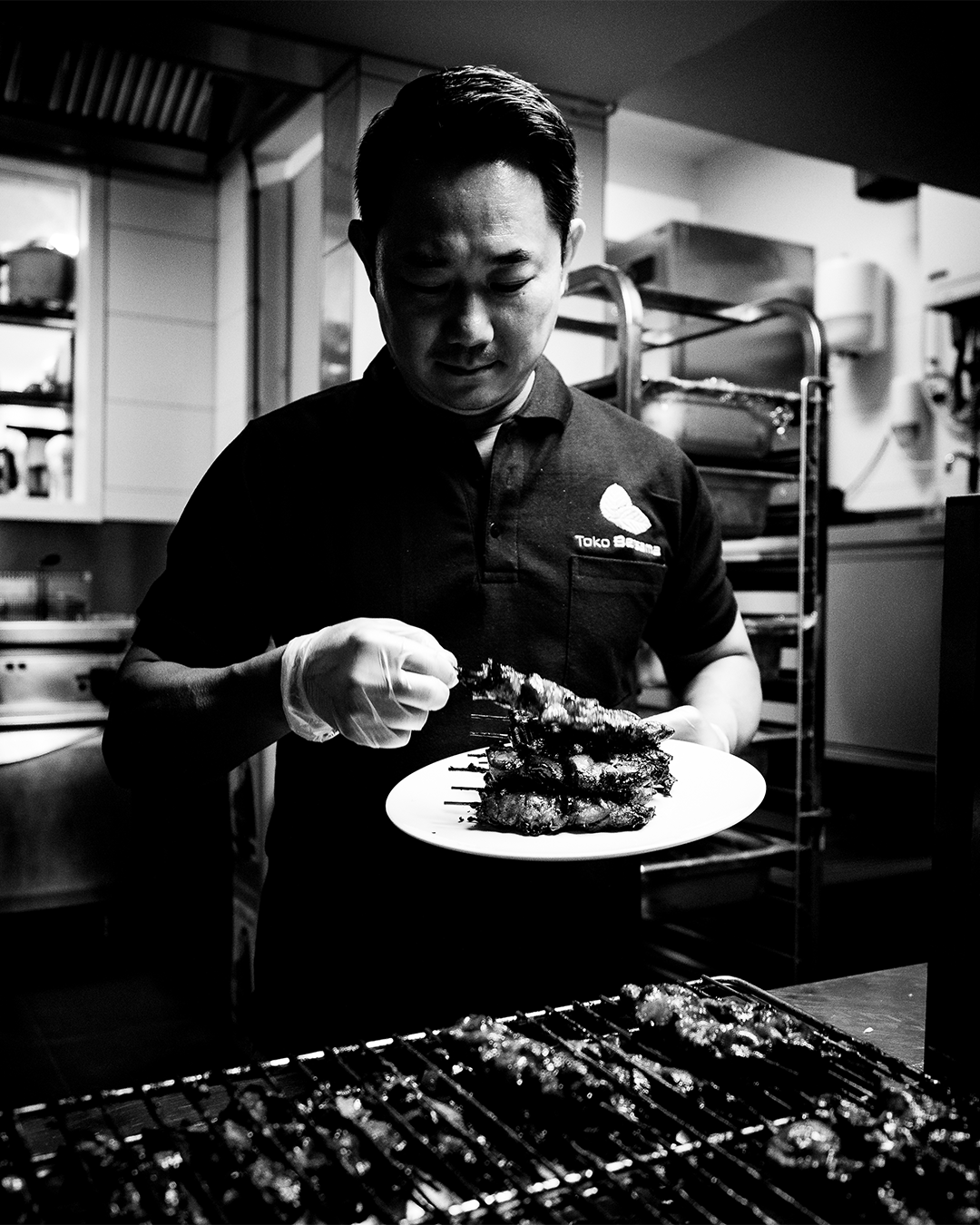 Chef in zwart overhemd met een bord met bereid voedsel; grillrooster op de voorgrond. Keukenomgeving.