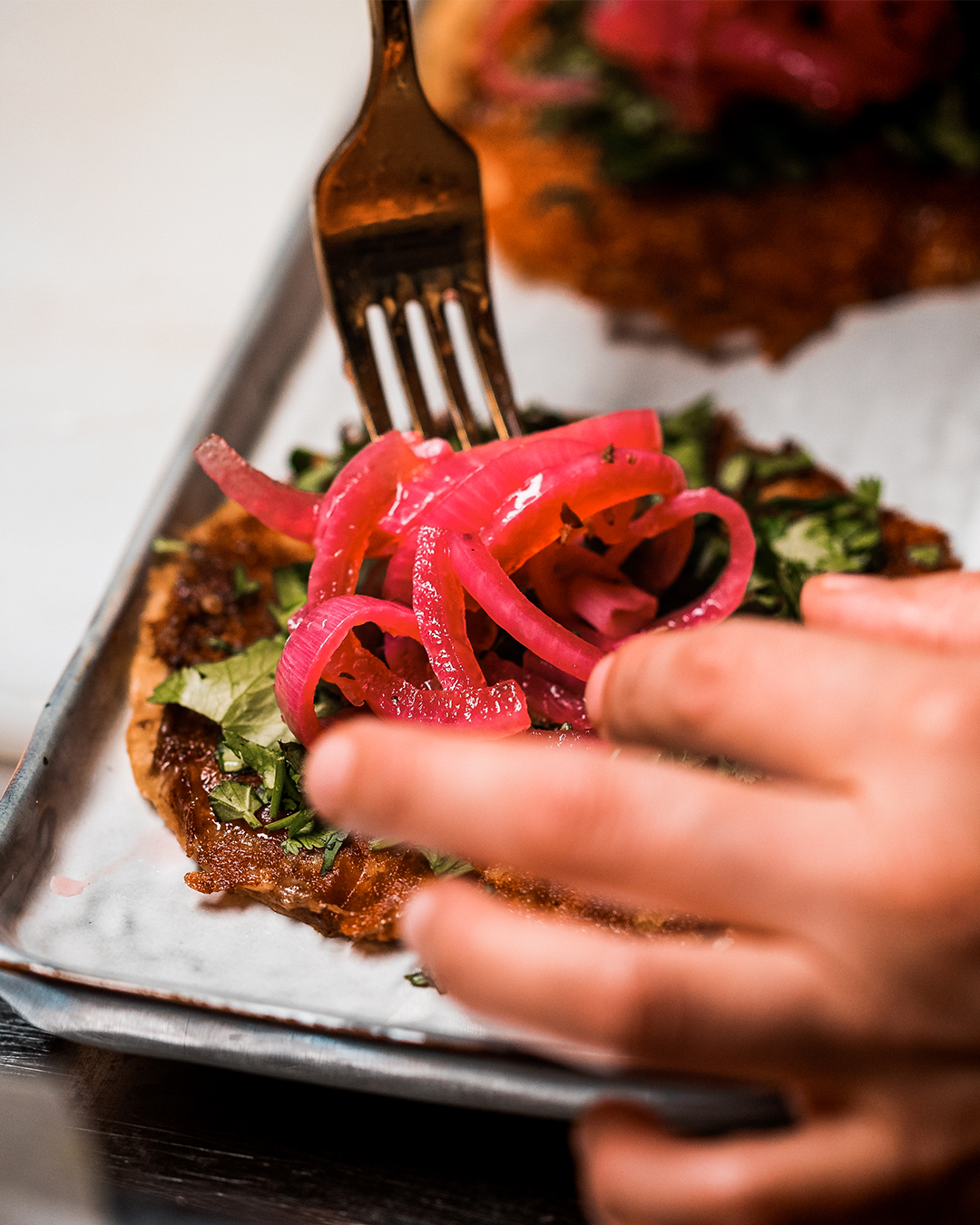 Met een vork voeg je ingelegde rode uien toe aan een klein gefrituurd broodje, belegd met groene bladgroenten.