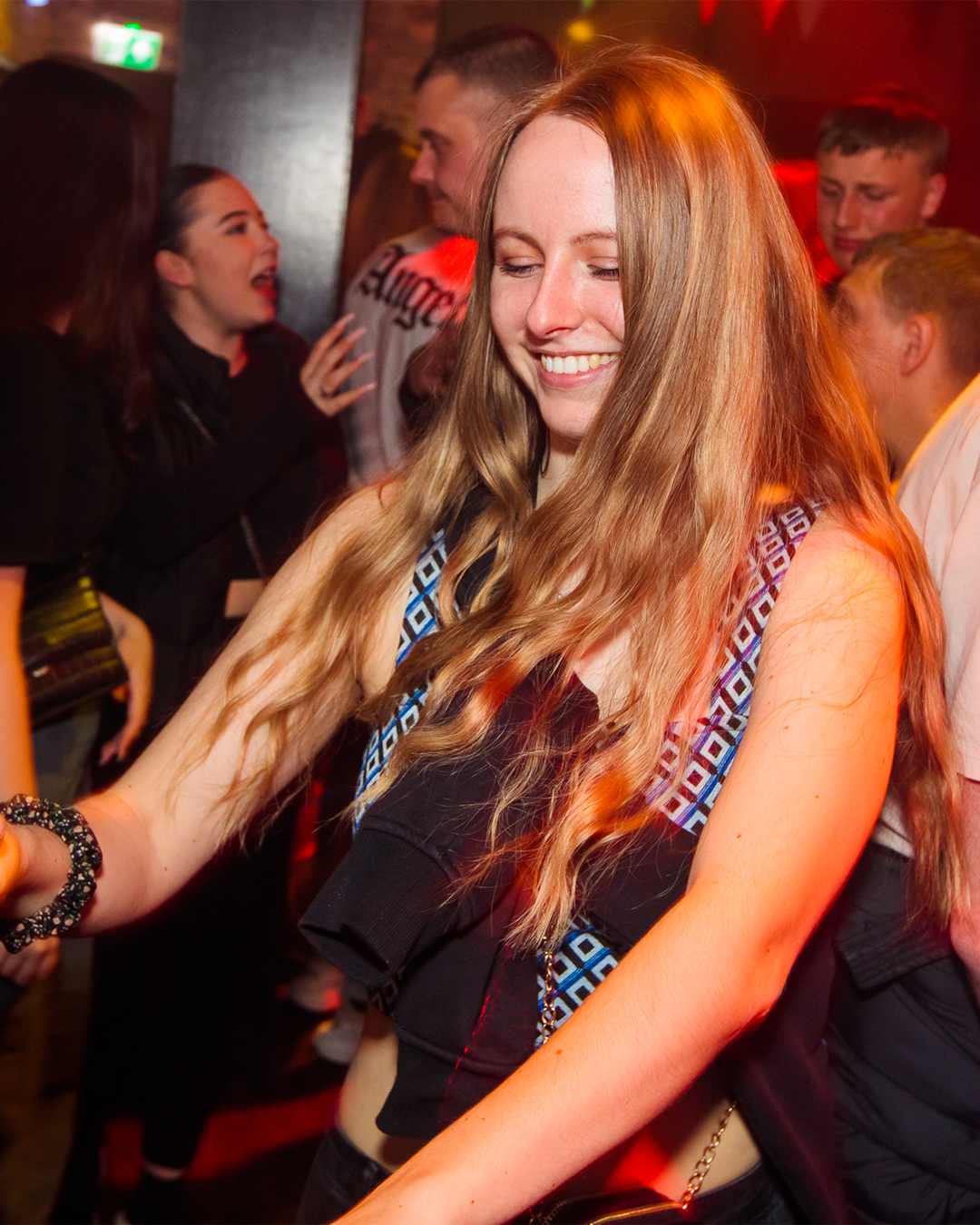 Een vrouw met lang blond haar lacht terwijl ze danst in een drukke club. Anderen op de achtergrond.