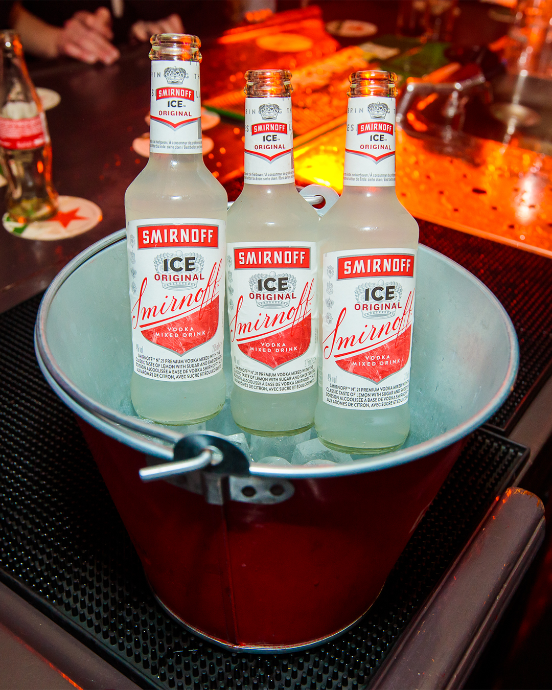 Drie flessen Smirnoff Ice in een rode ijsemmer op een bar, verlicht met oranje licht.