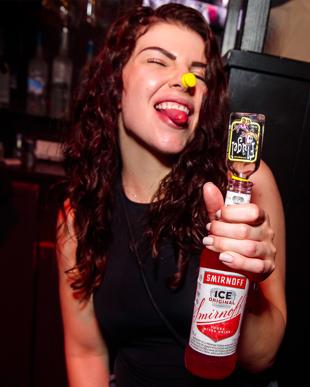 Een vrouw met een fles Smirnoff Ice in haar hand en een feestartikel op haar neus, steekt haar tong uit in een donkere bar.