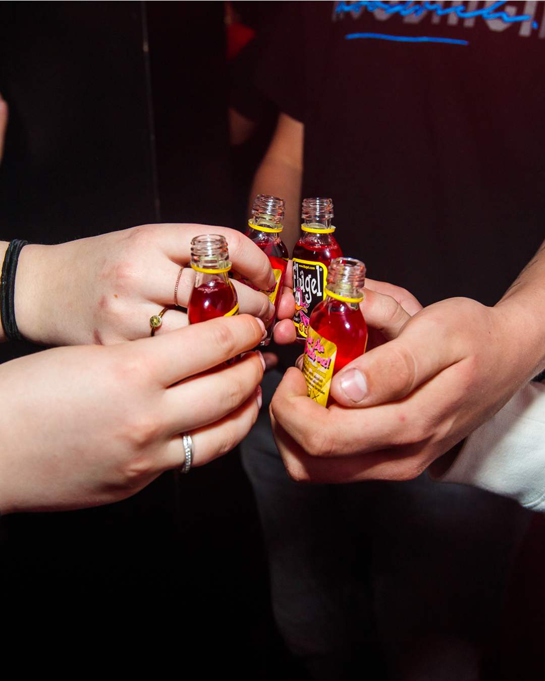 Vier mensen houden kleine flesjes met rode vloeistof omhoog, waarschijnlijk alcoholische shotjes, om een feestelijk toast uit te brengen.