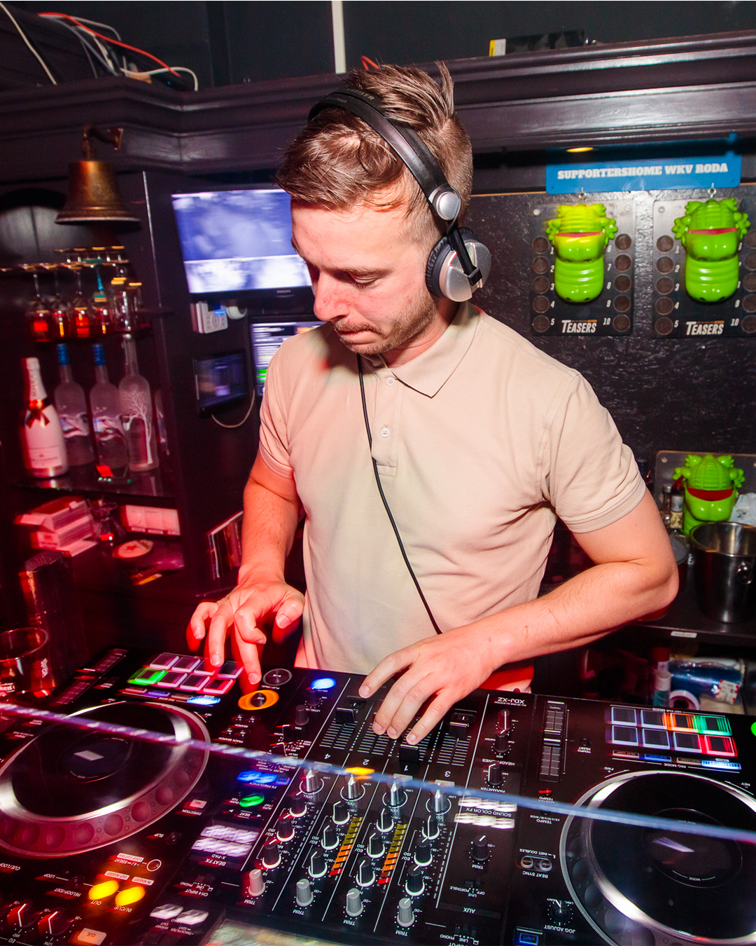 DJ in een club, die muziek mixt op een mengpaneel, met een koptelefoon op. Gedempt licht met flessen en neonreclames.