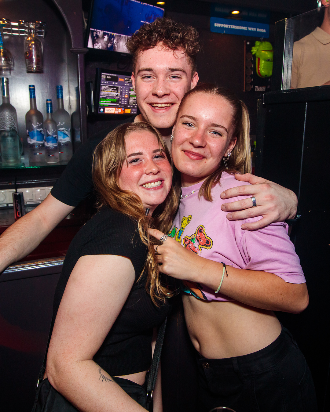 Drie lachende mensen poseren dicht bij elkaar in een schemerig verlichte bar; één van hen heeft zijn armen om de anderen heen geslagen.