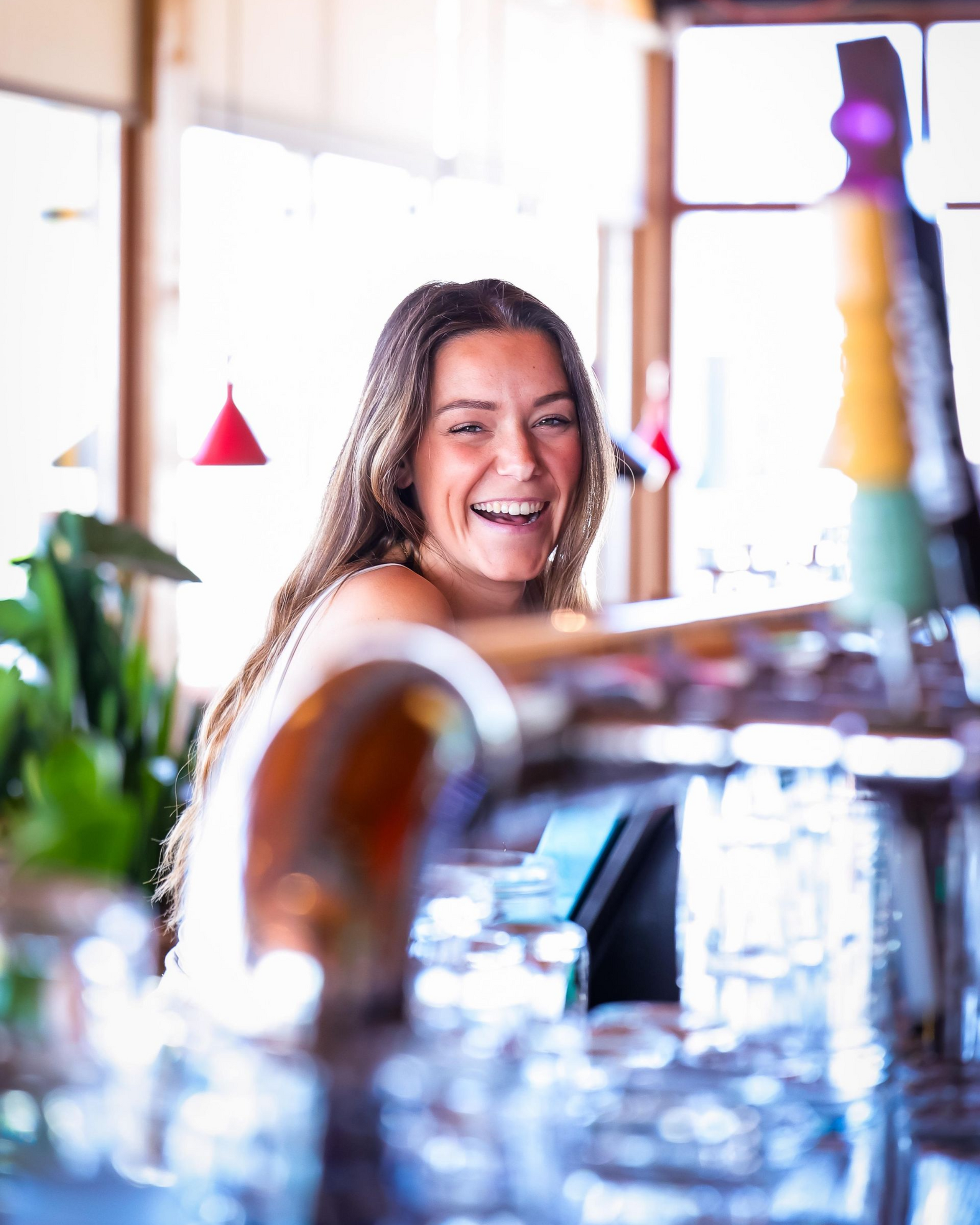Een vrouw lacht achter de bar, haar armen iets buiten beeld, met biertaps en glazen scherp in beeld.
