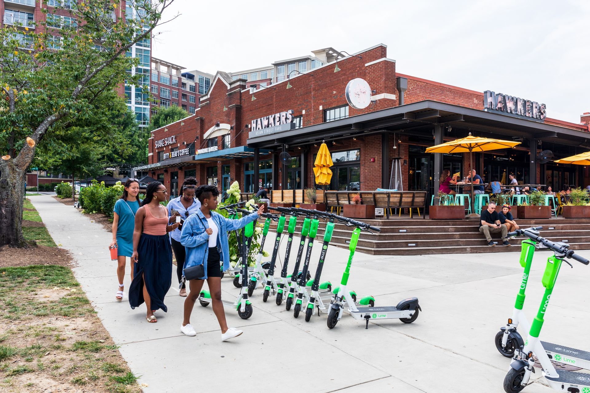People walking past parked green scooters on a sidewalk near brick building with outdoor seating in South End, Charlotte