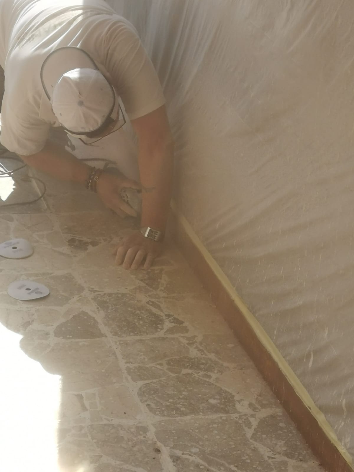 Person in a white shirt and cap sands a wooden trim along a wall, floor is marble-patterned.