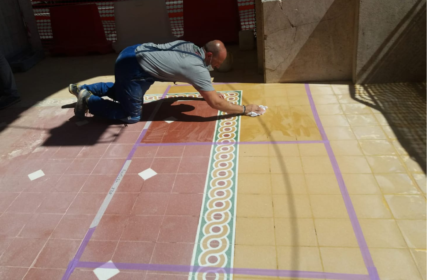 Man cleaning tiled floor, kneeling with cloth. Sunny outdoor setting, purple outline on tiles, red and yellow tiles.